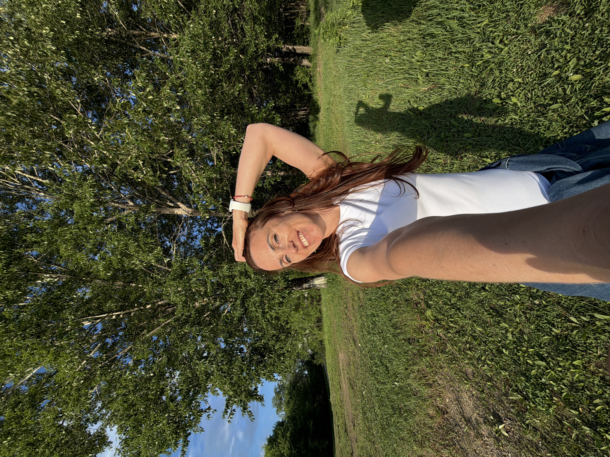 In Birobidzhan, Russia, a woman with long, brown hair and fair skin is taking a selfie outdoors. She is wearing a white t-shirt and denim shorts. Her right arm is raised and bent, with her hand resting on her head. She is smiling and looking at the camera. The background shows lush green grass and trees, with a bright blue sky above. The sun is casting a shadow of the woman on the ground.