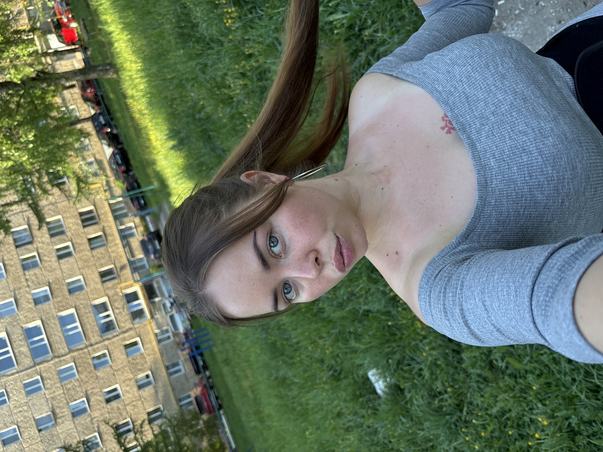 A fair-skinned woman with long brown hair and light-colored eyes takes a selfie in an outdoor setting. She wears a gray, long-sleeved shirt. A small red tattoo is visible on her upper chest. The background features a grassy area with yellow flowers, a building with many windows, and parked cars. The location is likely Izhevsk, Russia, based on the architectural style of the building.