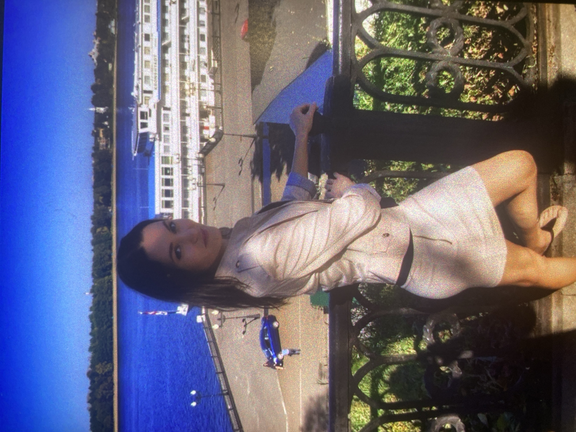 The image captures a woman posing on a sunny day in Saint Petersburg, Russia, with the Neva River as a prominent backdrop. The woman, positioned in the foreground, has long dark hair and is dressed in a light-colored jacket and a matching short skirt, paired with light flat shoes. She leans against an ornate dark metal railing, with one hand resting on it, and looks directly at the camera with a calm expression.
The bright sunlight suggests clear weather, casting distinct shadows from the railing and objects on the ground. Through the railing, green foliage is visible. Behind the woman, a wide, deep blue body of water, indicative of the Neva River, stretches into the distance. Several large white multi-decked ships are either docked or navigating the river, with the word "KRONSTADT" clearly visible on the side of one of the vessels. The far bank of the river features a dense treeline, with faint distant structures, possibly a church spire, visible beyond. A small blue car is parked on the paved promenade in the mid-ground, and lampposts line the waterfront.