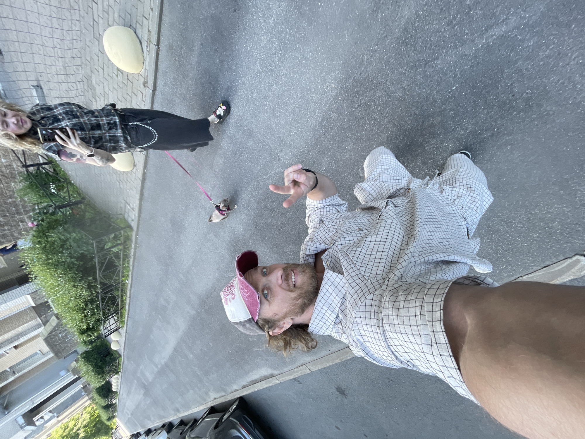 The image shows two people and a small dog on a paved area, possibly a sidewalk or street, in Saint Petersburg, Russia. The orientation of the image is landscape and sideways. The first subject, a man, is lying down and looking up at the camera, smiling. He is wearing a checkered shirt and a baseball cap. He is making a peace sign with his hand. The second subject, a woman, is standing and holding a leash attached to the small dog. She is wearing a plaid shirt and a long black skirt. She is holding a phone in her hand. The scene appears to be outdoors during the daytime, with sunlight visible. There are some green bushes and buildings in the background. The ground is made of asphalt and paving stones. No text is visible in the image.