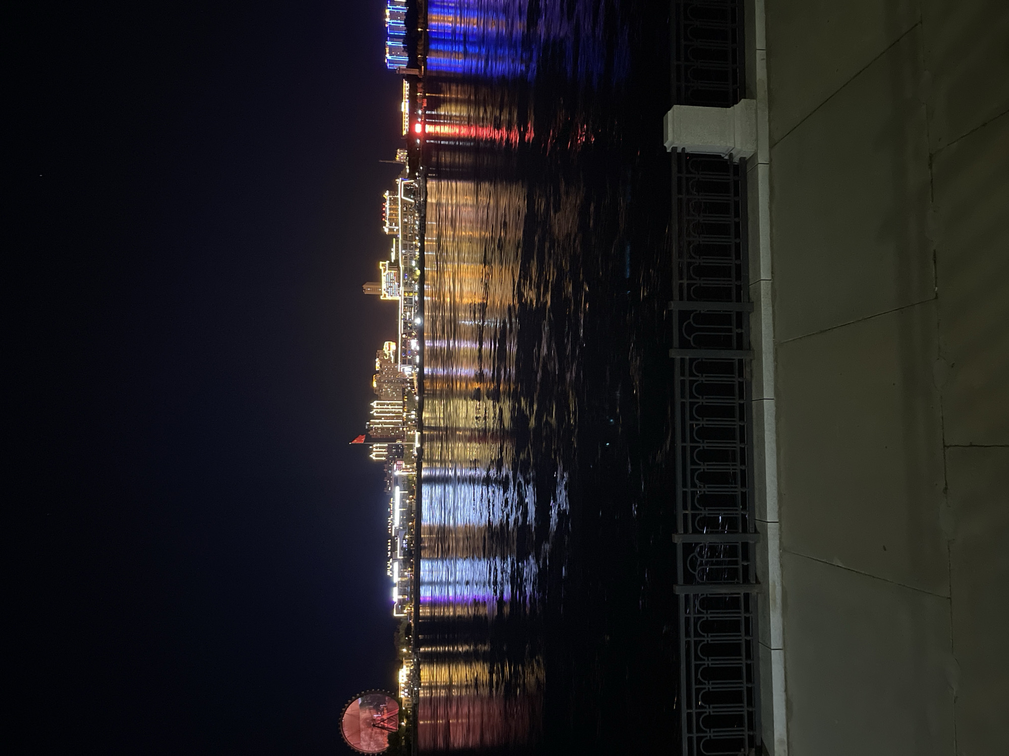 A night-time panoramic view of the Amur River embankment in Blagoveshchensk, Russia. The waterfront features a city skyline illuminated by numerous lights, casting reflections on the dark water. On the left, a Ferris wheel with red lights is visible. The buildings in the background are brightly lit with yellow, white, and blue lights. A red flag flies from a pole in front of one of the buildings. The foreground shows a paved walkway separated from the river by a decorative metal fence and a concrete pillar. The sky is dark, indicating it is night.