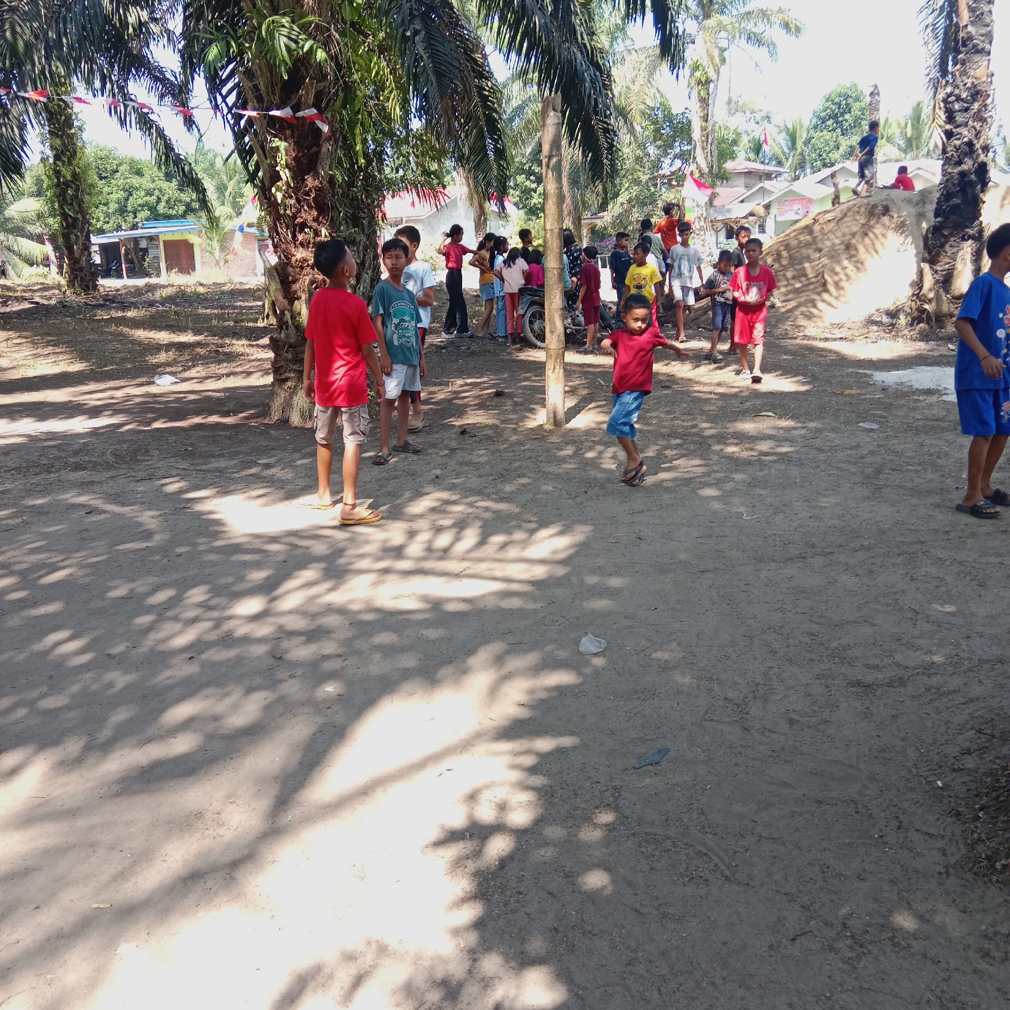 The image depicts an outdoor scene with a group of children in a sandy, dirt-covered area, likely a village or rural setting. Several palm trees with scattered Indonesian flags strung between them dominate the foreground, casting dappled shadows on the ground. In the background, several modest houses are visible, some with blue roofs, suggesting a residential area. A mound of earth, possibly a small hill, is present on the right side of the frame.
A young boy in a red t-shirt and blue shorts is in mid-air, appearing to jump or play. Several other children are standing and observing, some wearing casual clothing like t-shirts and shorts, and a few are wearing flip-flops. One boy in the foreground is wearing a red t-shirt and khaki shorts. Another boy to his left is wearing a green t-shirt and white shorts. There is a motorcycle parked in the middle ground, partially obscured by the children. The overall atmosphere appears to be one of a casual gathering or play in a warm, sunny climate.
Based on the context of palm trees, modest housing, and the overall environment, this scene is consistent with many rural or village areas in Dumai City, Indonesia, particularly in areas with agricultural land like palm oil plantations.