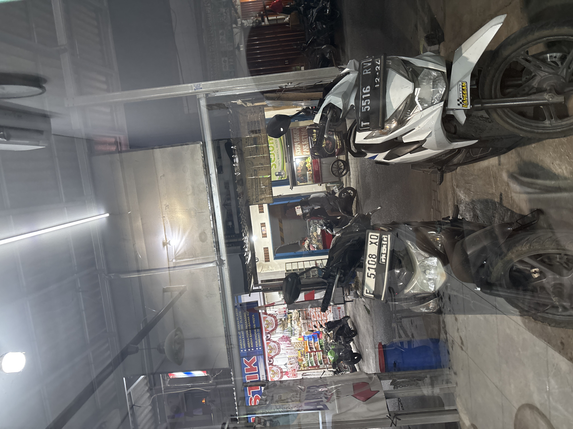 The image depicts a street scene in Wanajaya, Indonesia, likely at night given the artificial lighting. In the foreground, two motorcycles are parked. The motorcycle on the left is dark colored, with a license plate reading "F 5108 XQ". The motorcycle on the right is white and black, with a license plate reading "T 5516 RVI 12.24". Both motorcycles appear to be on a paved surface, possibly a sidewalk or street.
Behind the motorcycles, a storefront is visible. The storefront has a blue awning with text in Indonesian, including "ANIKA ALAT MAKAN PLASTIK" and other product-related phrases. Inside the store, shelves are stocked with various items, and some red and white banners are displayed.
Further down the street, other businesses are visible, including one with a sign that appears to advertise "AYAM BAKAR PAK RETNO". More parked motorcycles and a few individuals can be seen in the background, suggesting a commercial area. The overall impression is of a bustling street with small businesses and parked vehicles. The scene is viewed through a reflective surface, likely a window, which casts reflections on the image.