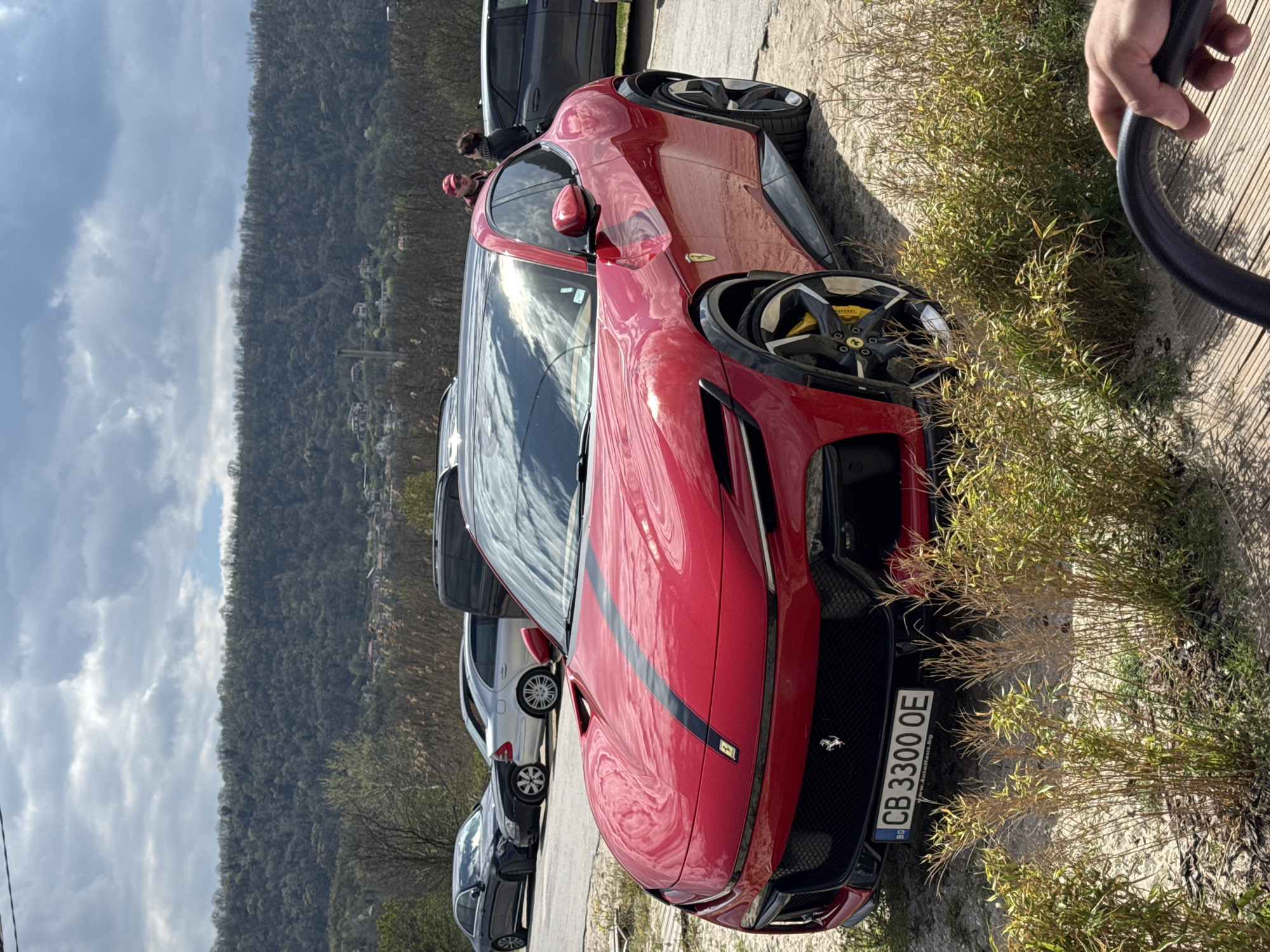 A red Ferrari with a black racing stripe is parked on a paved area next to tall, dry grass. The car's license plate reads "CB 3300 OE" and includes the Bulgarian flag. The background features a forested mountain range under a cloudy sky. Several other cars are parked in the background. Two people are partially visible near a dark grey vehicle. A black hose held by a hand appears on the right edge of the frame. This scene likely takes place in Bulgaria, judging by the license plate, possibly near Varna considering the license plate code.