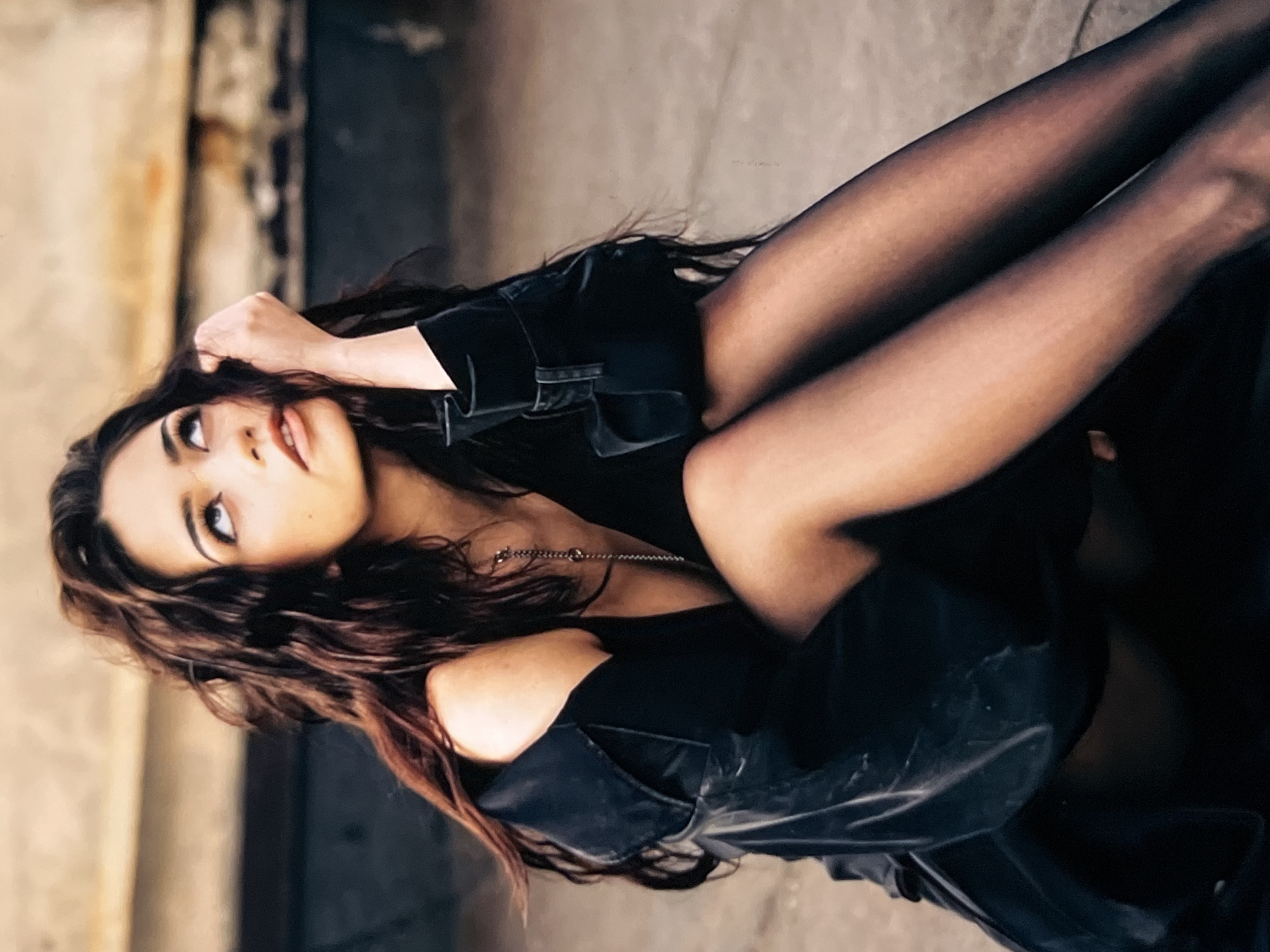 In a full-length, medium shot, a young woman with long, dark, wavy hair sits on a concrete surface outdoors, looking upwards. She is wearing a black off-the-shoulder jacket, a low-cut black top, and black sheer pantyhose. A silver chain necklace with small circular charms is visible around her neck. Her right hand is raised to her head, with her fingers gently touching her hair. The background consists of a blurred, weathered, urban-like structure, suggesting an outdoor setting in a city.