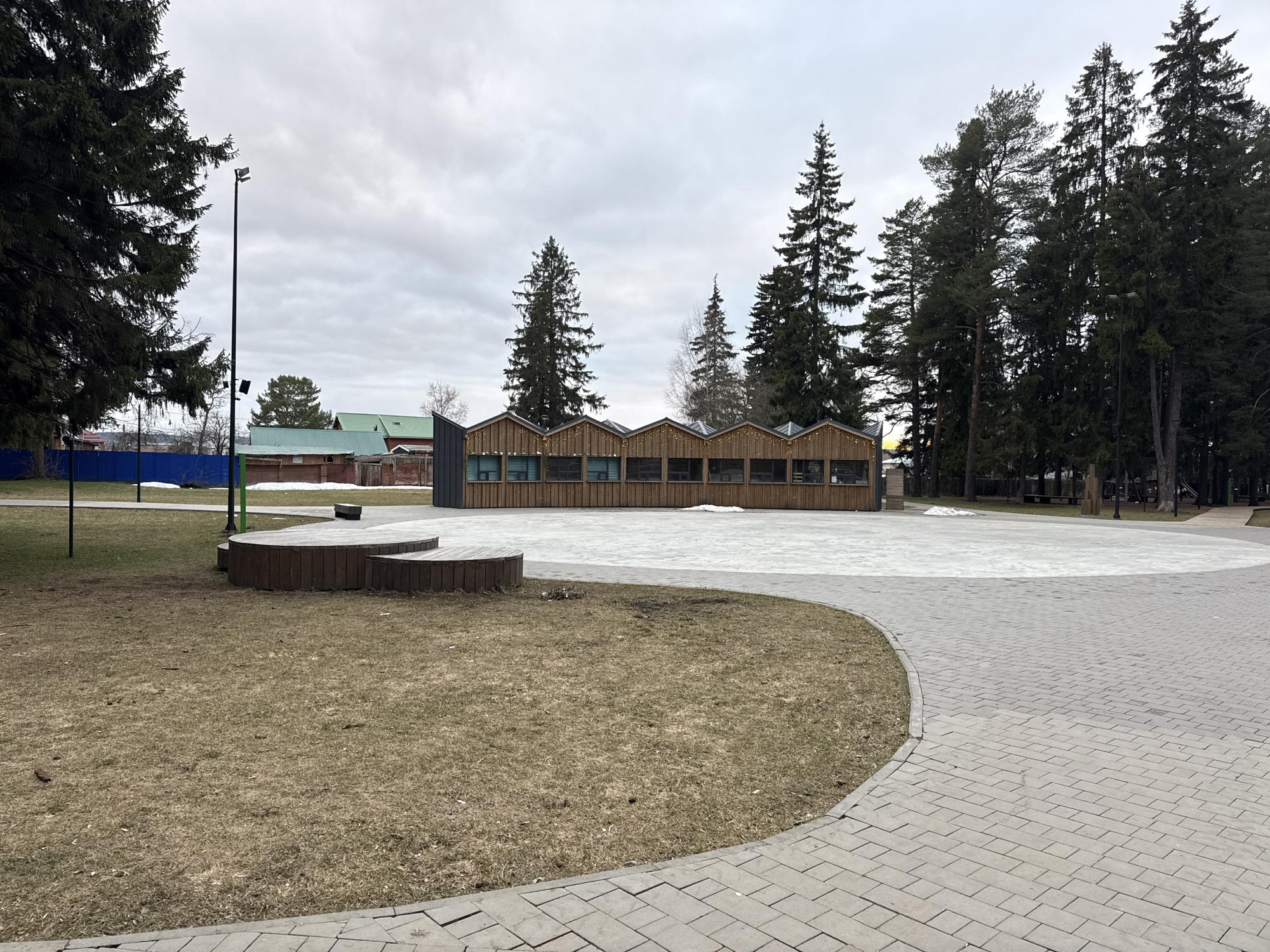 An outdoor scene in Votkinsk, Russia, depicts a public space on an overcast day, likely in late winter or early spring. The foreground features dry, light brown grass and a curved pathway made of light grey paving stones. To the left, a multi-tiered wooden seating structure or platform is visible on the grassy area.
In the mid-ground, a long, low-rise wooden building stands with a distinctive serrated or sawtooth roofline. The building has numerous rectangular windows along its facade, and small decorative string lights are visible along its roof edges, suggesting it may be a café, shop, or public facility. Beyond this building, a large paved area, possibly a square, is partially covered with patches of melting snow.
Tall coniferous trees, primarily pines and spruces, dominate the background and frame the scene on both sides, their dark green foliage contrasting with the muted sky. To the far left, a tall street light pole with floodlights extends upwards. In the distance, behind a blue corrugated fence, several smaller buildings with green roofs can be seen, hinting at a nearby residential area or village. The absence of people suggests a quiet moment in this public park or square. No discernible text is visible.