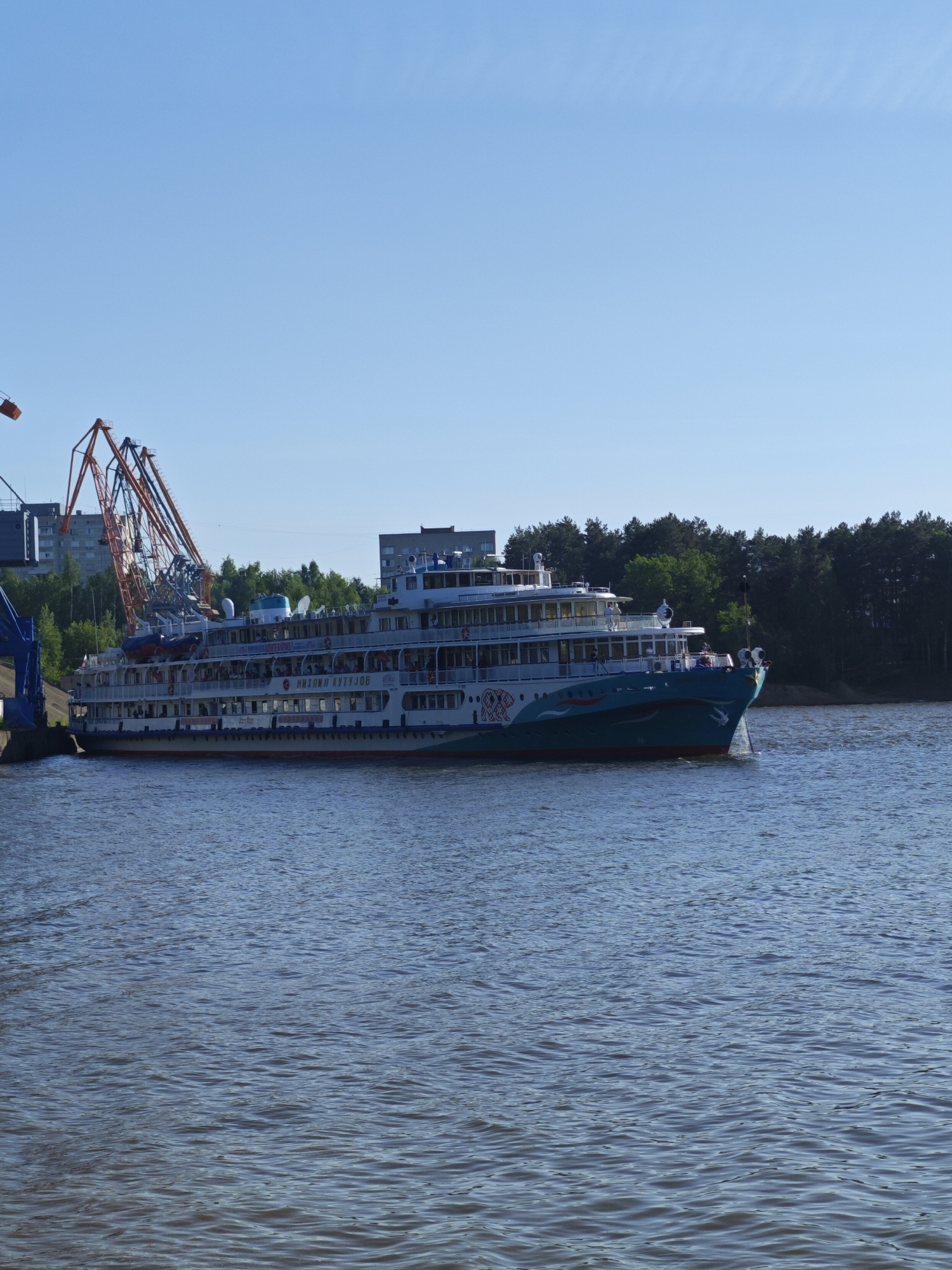 The image depicts a river scene in Chaikovsky, Russia. A large, multi-deck river cruise ship, with "Михаил Кутузов" (Mikhail Kutuzov) written on its side, is departing from a dock. The ship is painted in white, teal, and red, and has several rows of windows and balconies. Orange and blue cranes are visible next to the ship, suggesting an industrial or port area. Behind the cranes, there are residential buildings. Across the river, a line of trees indicates a wooded area. The water is calm, with visible ripples. The sky is clear and blue.