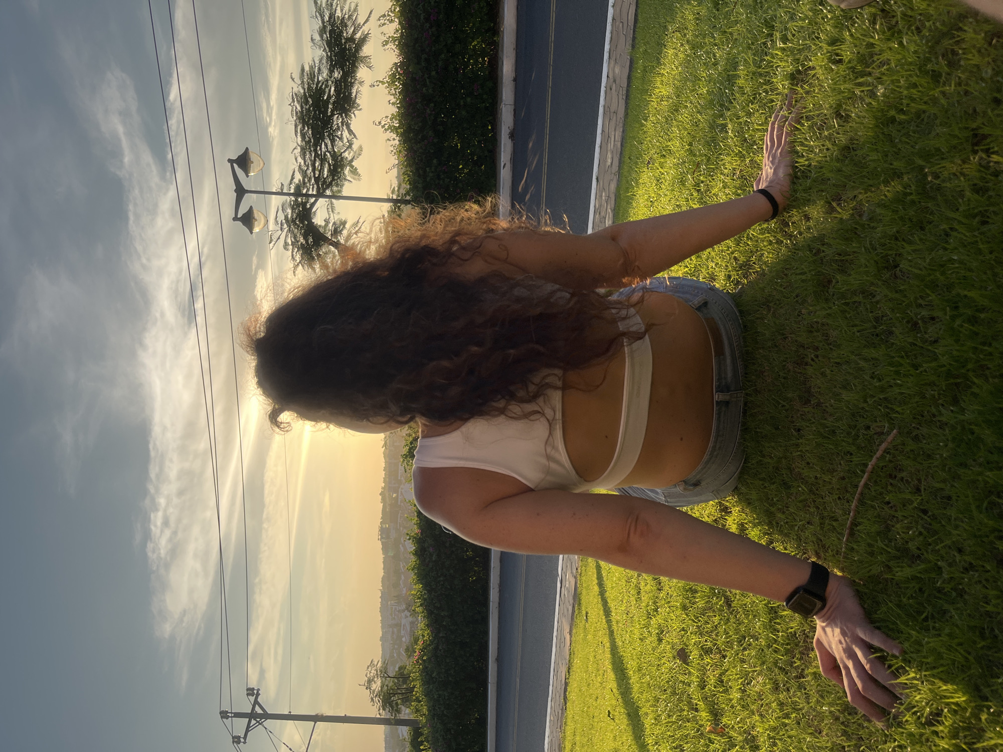 A woman with long, curly brown hair sits with her back to the camera on a grassy slope, facing a sunset over a city. She is wearing a white crop top and denim shorts. Her hands are resting on the grass on either side of her, and she has a watch on her left wrist. The scene appears to be in a park or a residential area. The sky is a pale blue with wispy clouds, and the sun is a bright orange orb near the horizon. In the background, there are trees, streetlights, and buildings. The grass is green and well-maintained, with shadows cast by the setting sun. The location is identified as Phu Thuy Ward, Vietnam.