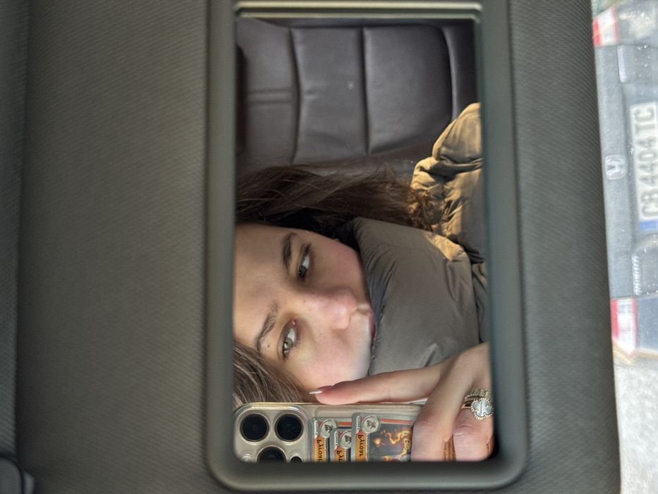 A woman is reflected in a car mirror. She is bundled in a grey jacket, and her brown hair is visible around her face. She holds a smartphone, the case adorned with various designs, and a ring is on her finger. The car seat is behind her, and a blurry image of a car number plate is visible outside the car window. The surrounding interior of the car is grey.