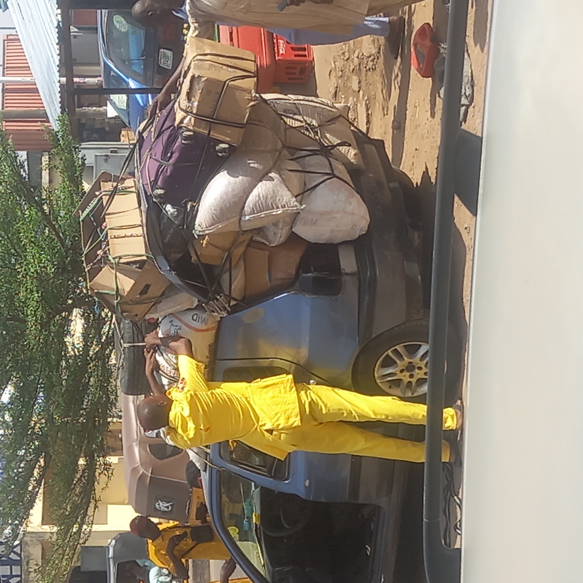 The image captures an outdoor scene, likely in Dadi Riba, Nigeria, depicting a heavily loaded dark-colored sedan or hatchback car parked on a dusty, sandy ground. A man dressed in a bright yellow full-body outfit is prominently featured, standing next to the car and actively securing a substantial amount of cargo piled on its roof.
The car's roof rack is overflowing with various items, including numerous cardboard boxes, several large white sacks (one of which clearly displays "MID" text), and what appears to be purple fabric or luggage. Black ropes or bungee cords are crisscrossing the cargo, tightly binding it to the vehicle. The man in yellow is shown holding onto some of these ropes, seemingly making final adjustments to the load.
Several other individuals are visible in the background and near the car, with at least one more person also appearing to wear a yellow uniform. In the background, structures with corrugated iron roofs and sections of red tile are visible, along with lush green foliage from a tree. A blue car is partially seen further back. Near the rear of the heavily loaded car, red plastic crates, possibly beverage containers, are stacked. A single red plastic bucket or container lies on the ground in the mid-ground.
The scene takes place during daytime under bright sunlight, evidenced by the clear shadows cast on the ground. The overall impression is of a busy transport or marketplace environment, characterized by diligent manual labor and preparation for the movement of goods.