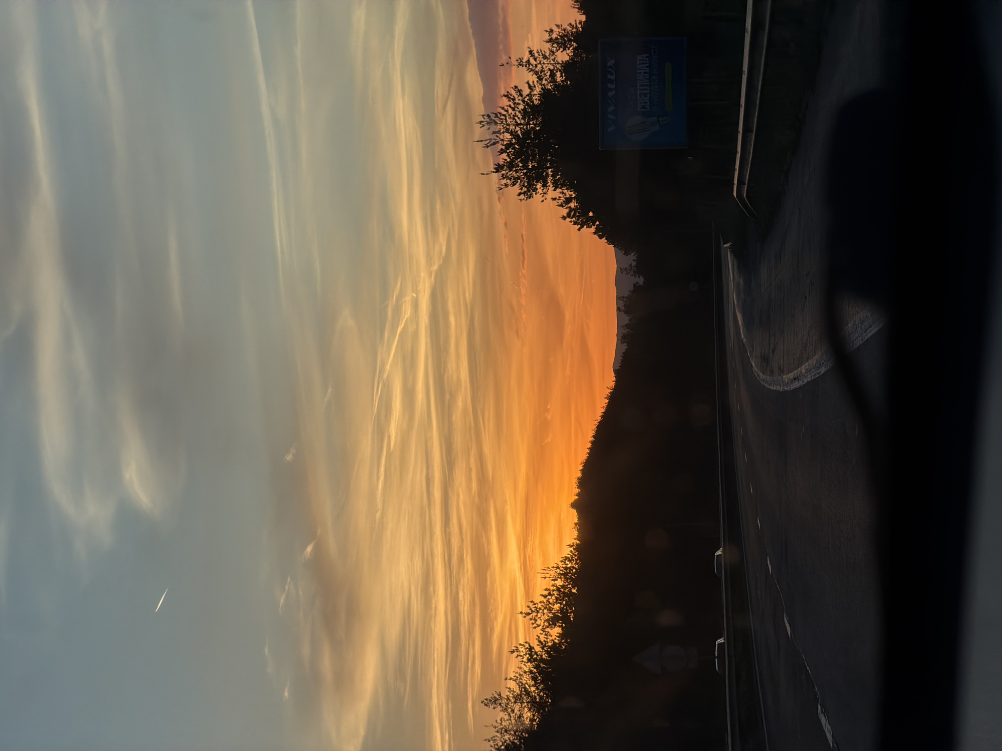 The image shows a sunset sky with streaks of orange and yellow clouds. The scene includes a road with a barrier and a sign in Bulgarian that reads "Vivalux" and "Светлината е здраве и живот" (The light is health and life). The location is likely near Dolna Vasilitsa, Bulgaria. There is a silhouette of trees and foliage along the horizon. A possible contrail from a plane can be seen in the left side of the sky. The photograph appears to be taken from inside a vehicle.
