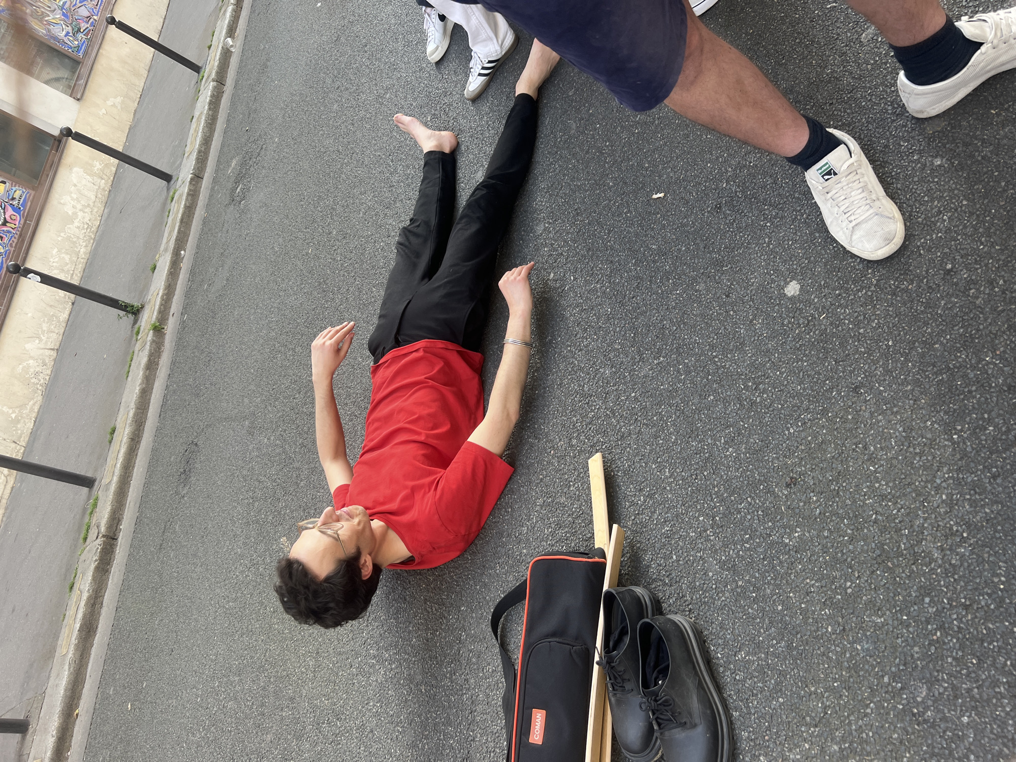 The image depicts a man lying on an asphalt street in Paris, France. He is wearing black pants and a red t-shirt. He has bare feet, except for one foot near a person standing over him. His hands are outstretched. To his side is a black bag, a pair of black shoes, and a wooden object that may be part of an easel. Above him, two individuals are standing. One is barefoot, and the other is wearing white sneakers and navy socks. The street is next to a curb and a building with windows.