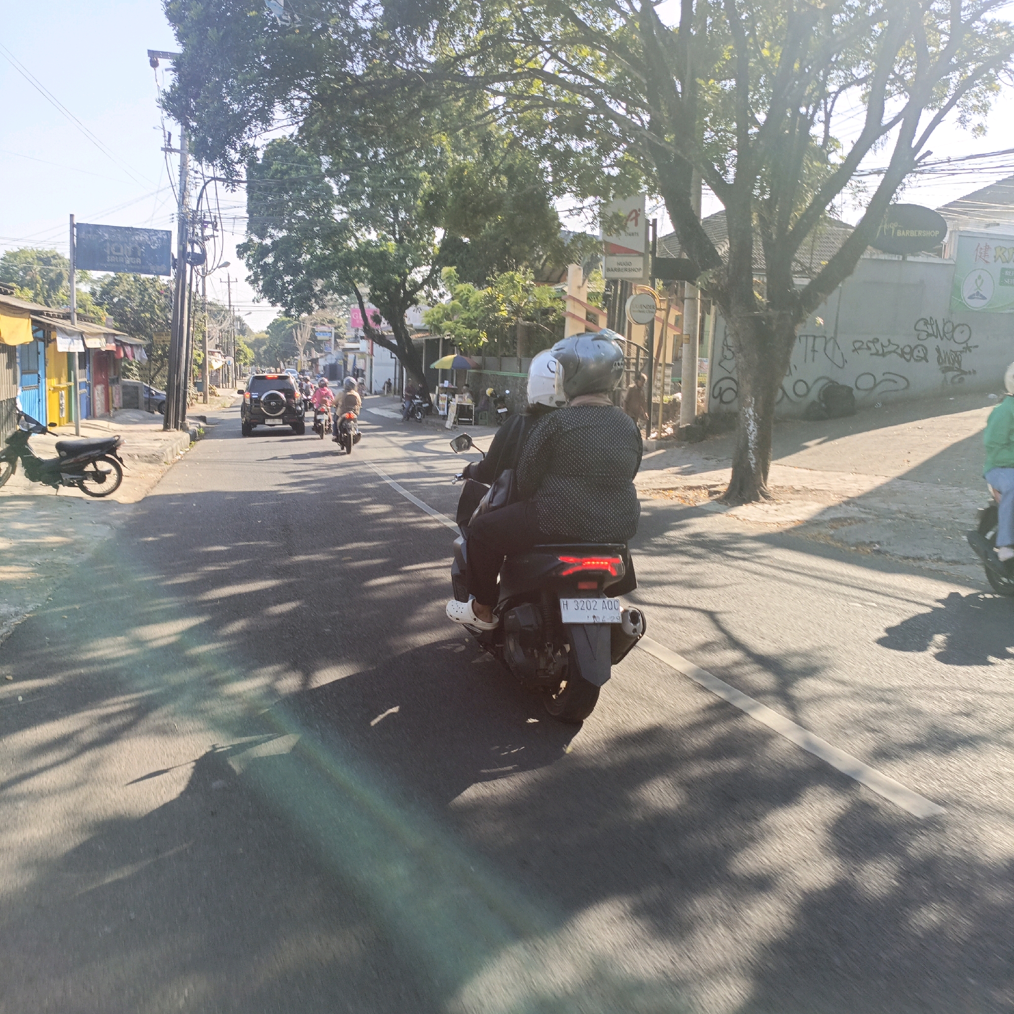 This image captures a street scene in Salatiga, Indonesia, during daylight. The perspective is from behind a motorcycle carrying two individuals, both wearing helmets. The motorcycle, with license plate H 3202 AOG, is in motion on an asphalt road. To the left of the road, there are small shops with colorful facades and laundry hanging outside. Across the street, on the right, is a wall with graffiti, a tree casting shadows, and signs indicating businesses such as "HUGO BARBERSHOP." Several other motorcycles and a dark SUV are visible further down the street, suggesting moderate traffic. The overall environment appears to be a typical urban street with a mix of commercial establishments and residential buildings. The sunlight creates strong shadows of trees on the road, indicating a sunny day.