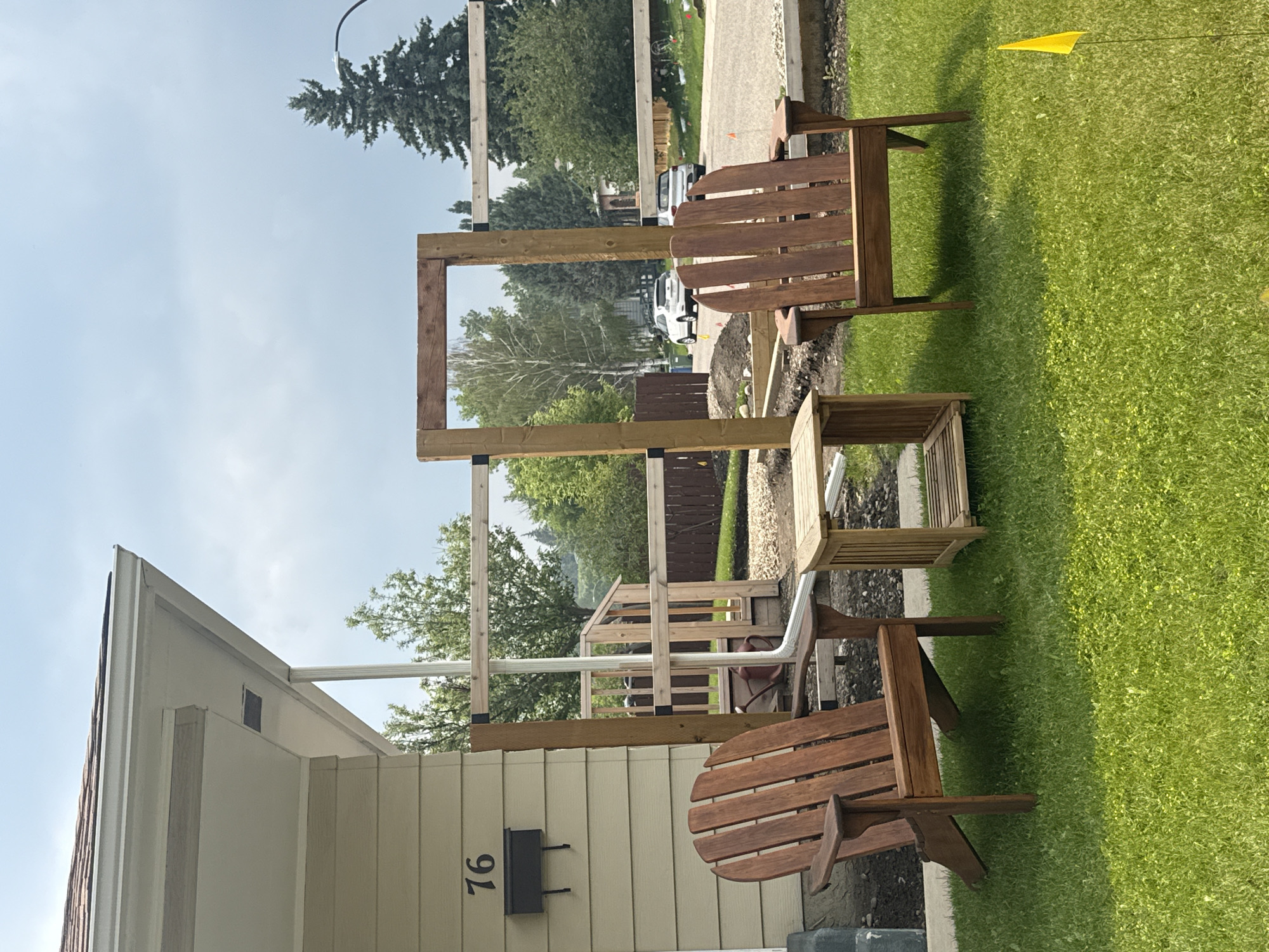 The image shows a residential setting, likely a backyard in Calgary, Canada. The scene is dominated by a partially constructed wooden pergola or trellis structure in the center. Two wooden Adirondack chairs and a small wooden side table are arranged on a green lawn in front of the structure.
To the left of the pergola is a portion of a light-colored house with a dark mailbox affixed to the siding, displaying the number "76". A downspout runs along the side of the house. Behind the house and pergola, a paved driveway is visible with two white cars parked on it. Further in the background, there are trees, fences, and other residential properties, indicating a suburban neighborhood.
The time of day appears to be daytime, with a clear or partly cloudy sky. The lighting suggests it's not early morning or late evening. The weather seems pleasant, suitable for outdoor activities, given the presence of the chairs and the well-maintained lawn. A small yellow flag is visible on a pole in the grass to the right of the scene.
There are no people visible in the image. The primary objects are the wooden furniture, the unfinished wooden structure, and the house. The overall impression is one of a home improvement project underway in a typical suburban environment. The scene is static, with no discernible activity or event taking place beyond the construction. The visible text is limited to the house number "76".