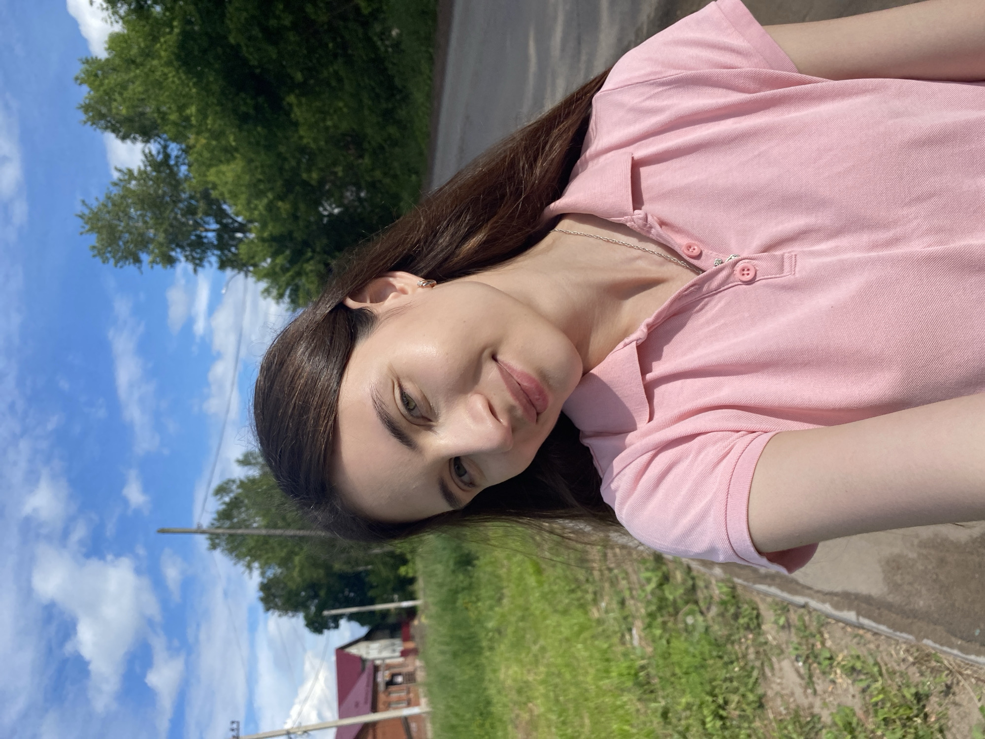 The image is a selfie taken outdoors. A young woman with long, dark brown hair and light skin is in the foreground, wearing a pink polo shirt and a silver necklace. She is smiling at the camera. The background shows a rural or suburban scene with a dirt road, a grassy area, trees, and houses with red roofs. The sky is blue with some white clouds. The location is identified as Votkinsk, Russia.