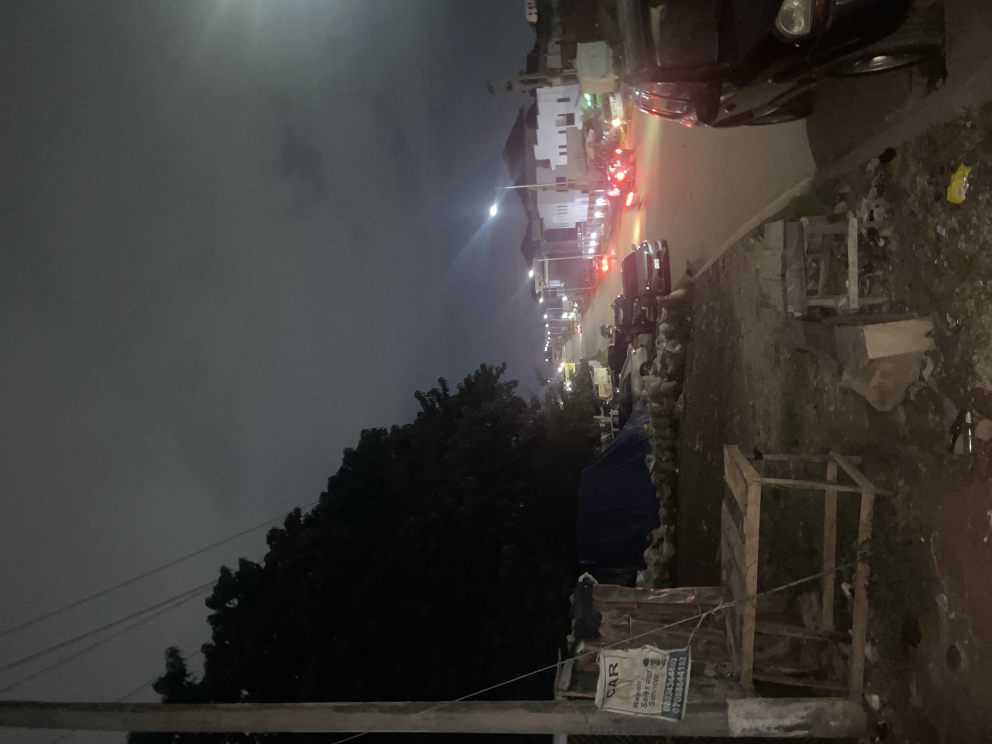 The image depicts a dimly lit street scene at night in Guzape, Nigeria. A prominent utility pole stands to the left, with power lines stretching across the dark sky. Adjacent to the pole, there is a makeshift stall or structure with a sign advertising "CAR Repair, Sales and Services" and two phone numbers. To the right of this stall, a blue tarp is visible, possibly covering goods or equipment. Further down the street, cars are driving, their headlights and taillights creating streaks of light against the darkness. Modern buildings with illuminated windows line the street, suggesting a developed urban area. The overall impression is of a residential or commercial street at night, with evidence of local businesses and ongoing traffic.