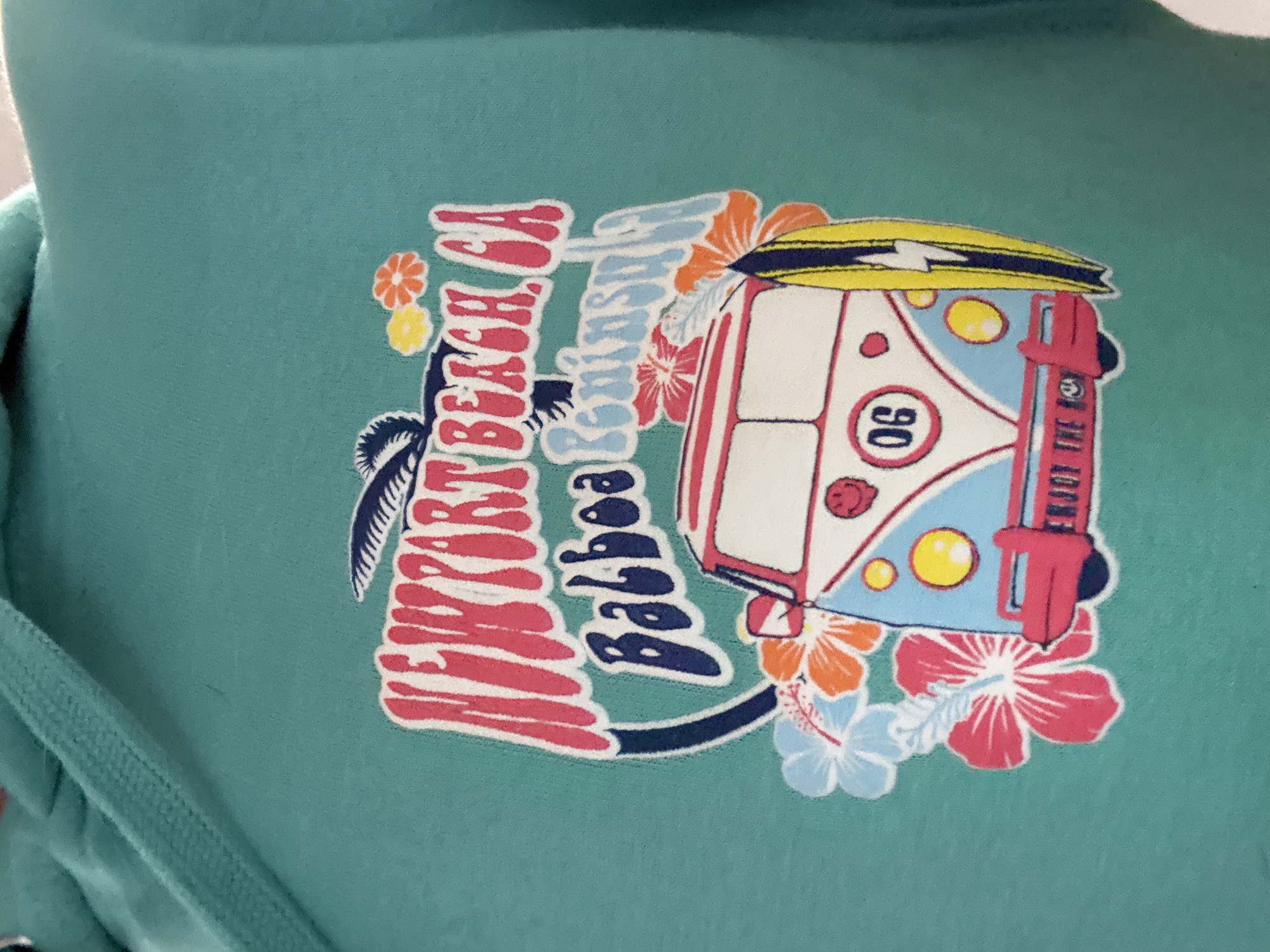 The image displays a close-up of a teal-colored garment, possibly a hoodie or sweatshirt, featuring a vibrant graphic design. The fabric has a soft, textured appearance.
The central subject of the graphic is a stylized vintage surf van, reminiscent of a Volkswagen Microbus, viewed from its front-right corner. The van is predominantly white and light blue, with red outlines and accents, including red taillights and what appears to be a red bumper or frame around the front. A yellow surfboard with a black stripe and white lightning bolt design is strapped to the roof. The front of the van has two round yellow headlights and a circular white emblem containing the number "06" in black. On the rear of the van, partially visible text reads "ENJOY THE RIDE".
Surrounding the van and integrated into the design are elements evoking a tropical, beach-side atmosphere. Large, bold text in red with a white outline reads "NEWPORT BEACH, CA", stacked vertically. Below this, in a light blue script with a white outline, is the word "Balboa". A dark silhouette of a palm tree is visible to the left of the text. Various tropical flowers in shades of orange, pink, yellow, and light blue are scattered around the graphic, adding to the colorful, summer-like feel.
The overall scene depicted by the graphic suggests a relaxed, Californian surf culture, associated with the iconic coastal locations of Newport Beach and Balboa. No specific activity is taking place, but the imagery evokes a sense of leisure, travel, and enjoying the outdoors.