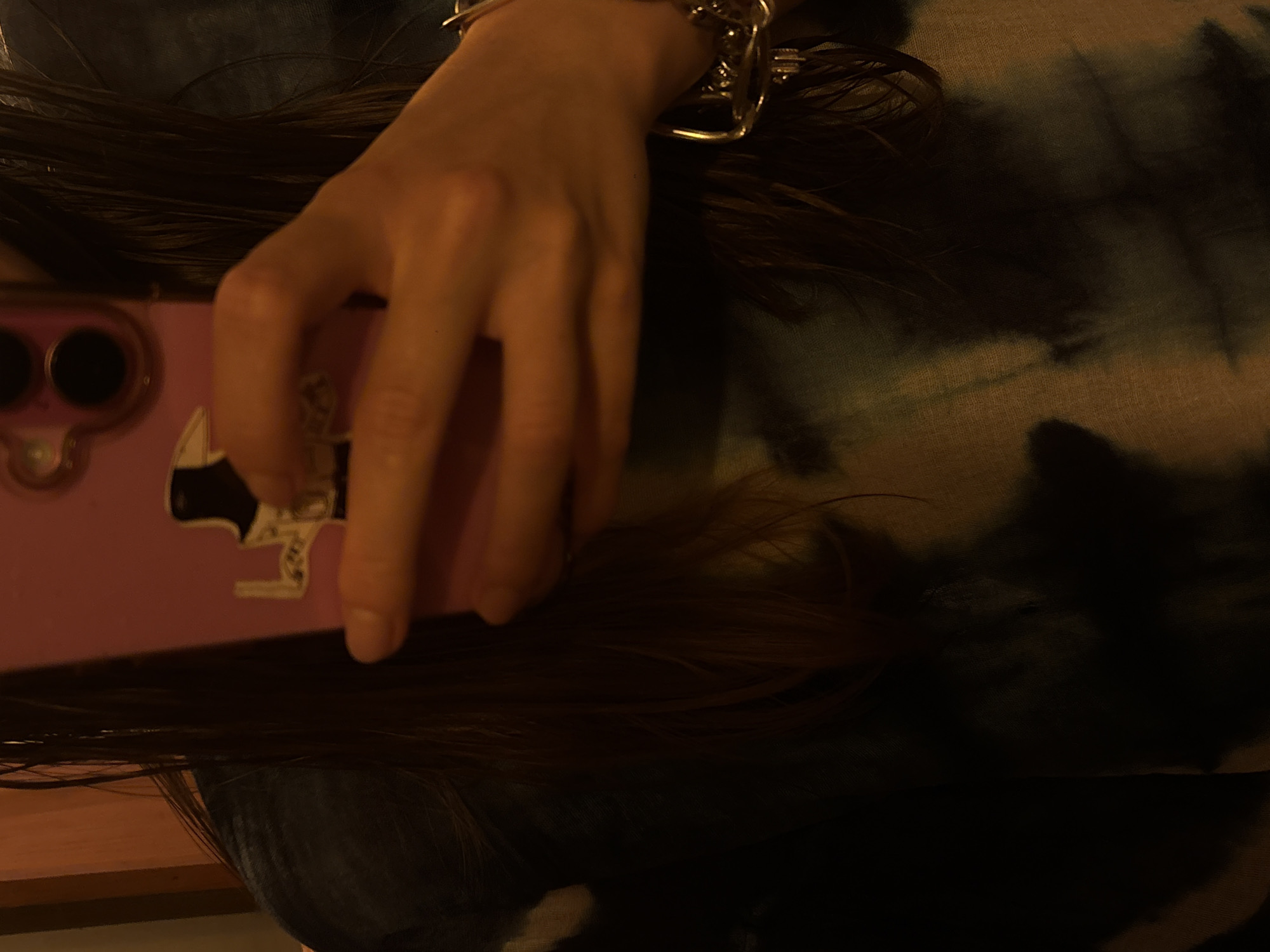 This close-up image, captured in Mexico City, Mexico, features a person's hand holding a pink smartphone. The hand, positioned centrally, has a metallic bracelet on the wrist. Long, dark hair surrounds the hand and phone. The pink phone case displays a sticker of a black handgun. The multi-lens camera module of the smartphone is partially visible on the left side of the frame. The background consists of a dark, tie-dye patterned fabric, likely clothing, and a hint of a wooden surface is visible in the lower left corner. The lighting is dim, suggesting an indoor setting during evening or nighttime. The primary activity depicted is the person holding the phone. No discernible text is visible within the frame.