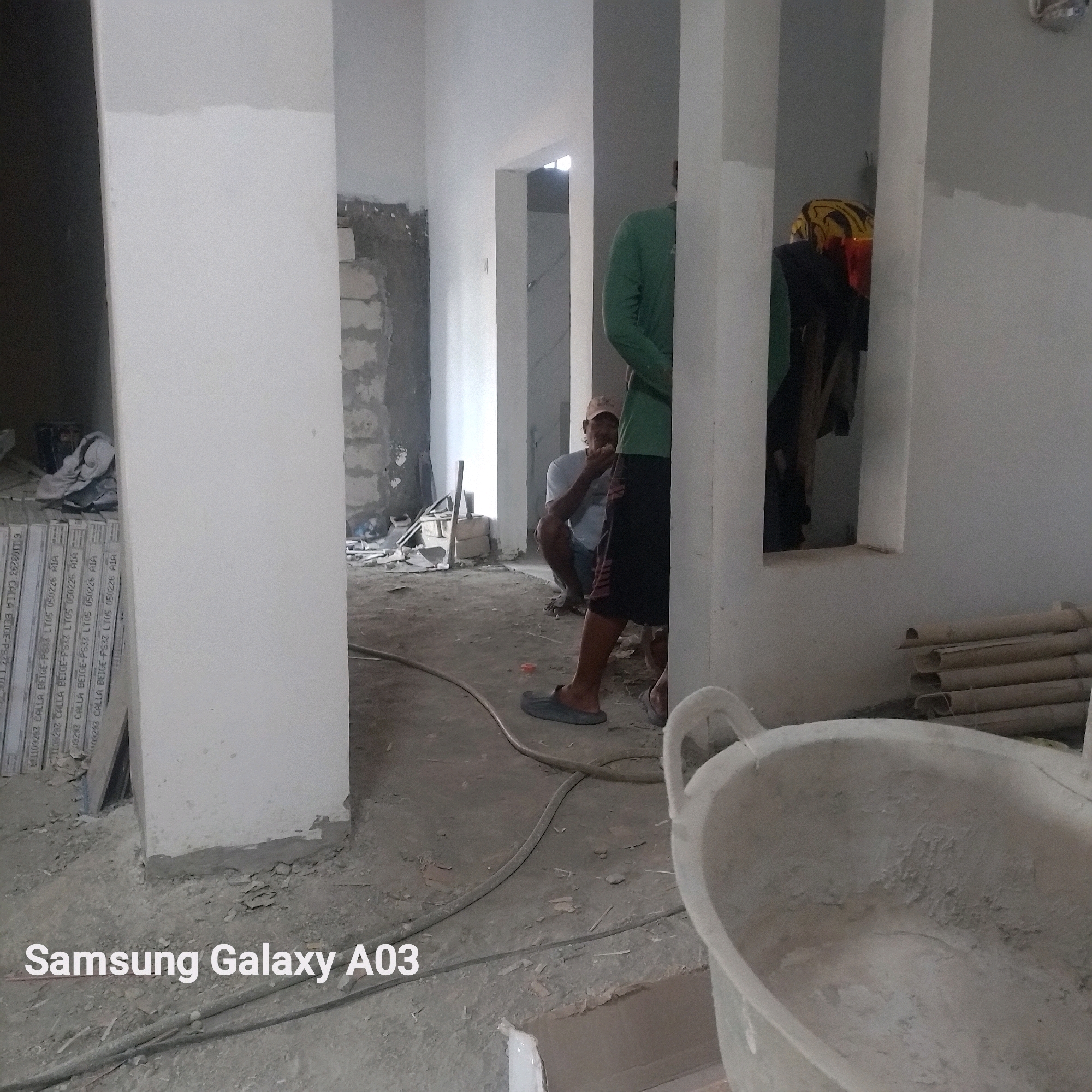 This image depicts an indoor construction site, likely a residential property undergoing renovation or building. The scene is characterized by unfinished walls, exposed building materials such as cement blocks, and a general state of disarray common in such environments. There are at least two individuals visible; one is partially obscured, wearing a green long-sleeved shirt and dark shorts, and appears to be walking. Another person, wearing a light-colored t-shirt and a baseball cap, is squatting and holding something to their mouth, possibly eating.
The environment is dusty with concrete debris scattered on the floor, along with construction tools and materials. A large plastic tub, possibly for mixing cement or carrying debris, is prominently featured in the foreground. A stack of what appear to be construction boards or tiles is visible on the left side of the image, with legible text indicating they are "CALLA BEIGE-PS33." A hose runs across the floor, suggesting plumbing or electrical work is in progress. The lighting appears to be natural, coming from openings in the structure.
The presence of construction work and the overall unfinished state of the interior suggest a building or renovation project. The text on the construction materials may offer clues to the specific type of material being used. The watermark "Samsung Galaxy A03" indicates the device used to capture the image. While the image itself does not contain any direct visual cues to identify the specific location within Indramayu, Indonesia, the context of a construction site is clear.