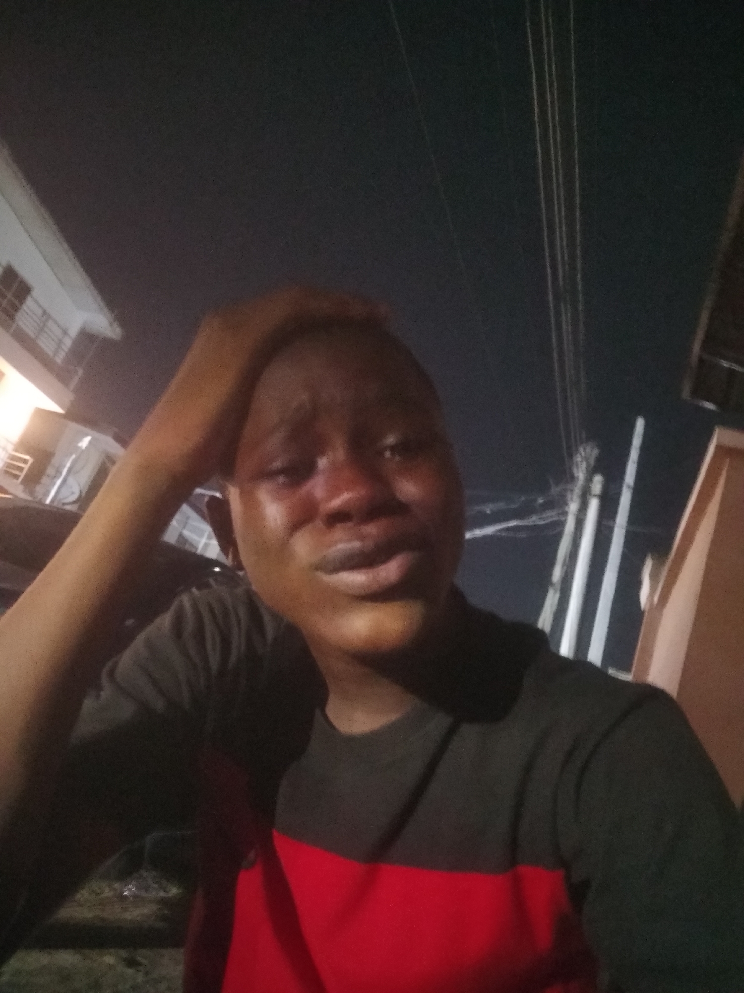 This is a close-up, dimly lit, nighttime portrait or selfie featuring a young person, likely male, of dark complexion. The subject is positioned centrally, looking towards the viewer, and exhibiting clear signs of distress. Their eyes are red and watery, suggesting they are crying, and their mouth is slightly open. Their right hand is raised and resting on their forehead or head, a gesture commonly associated with sadness, despair, or contemplation. The subject is wearing a t-shirt that is dark grey or black on the top half and red on the bottom half, with a horizontal seam across the chest.
The background elements indicate an outdoor, urban, or suburban setting. To the left, part of a multi-story building with illuminated windows and railings is visible. To the right, several utility poles or structures with overhead wires stand against the dark night sky. A portion of a light-colored building or wall is also visible on the far right. The overall lighting is artificial and sparse, highlighting the subject's face while keeping the background largely in shadow. The image captures a moment of raw emotion, possibly during a difficult situation. No text is visible in the image. The location context is Shangisha, Nigeria.