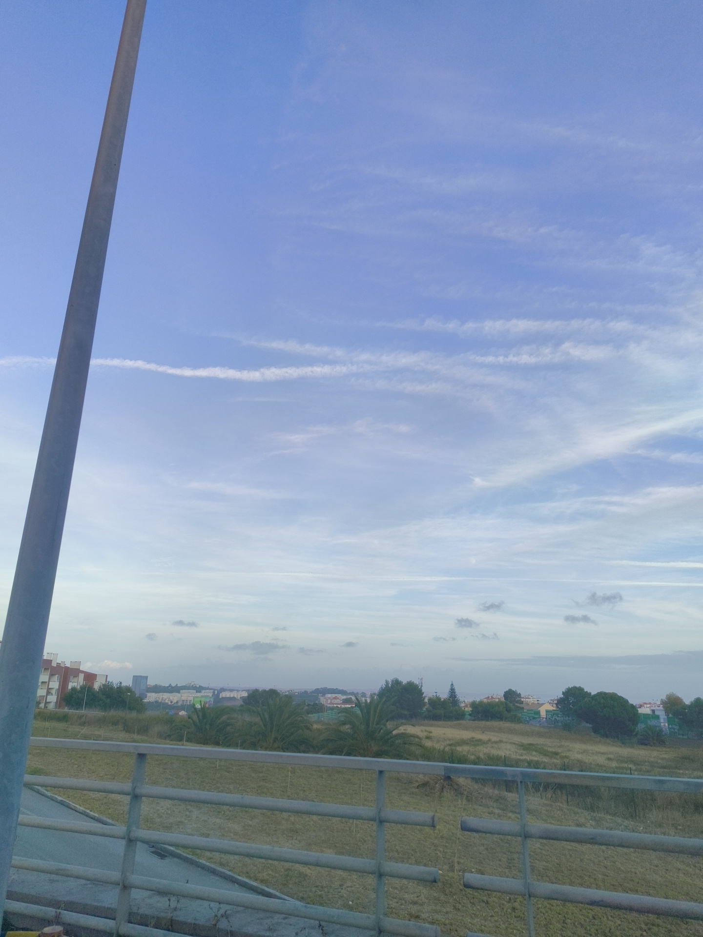 The image depicts an outdoor scene with a clear blue sky and wispy white clouds. In the foreground, a grey utility pole is visible on the left, extending from the bottom to the top of the frame. A metal railing with horizontal bars runs across the bottom third of the image, with a grey paved walkway behind it. The background shows a landscape with dry grass, scattered trees including palm trees, and buildings in the distance. The buildings suggest an urban or suburban environment. The scene appears to be located in Cascais, Portugal, given the context provided. There are no people or animals visible in the image, and no specific actions or interactions are taking place. The overall impression is a view of a town or city from a slightly elevated position, possibly a bridge or walkway, on a day with fair weather.