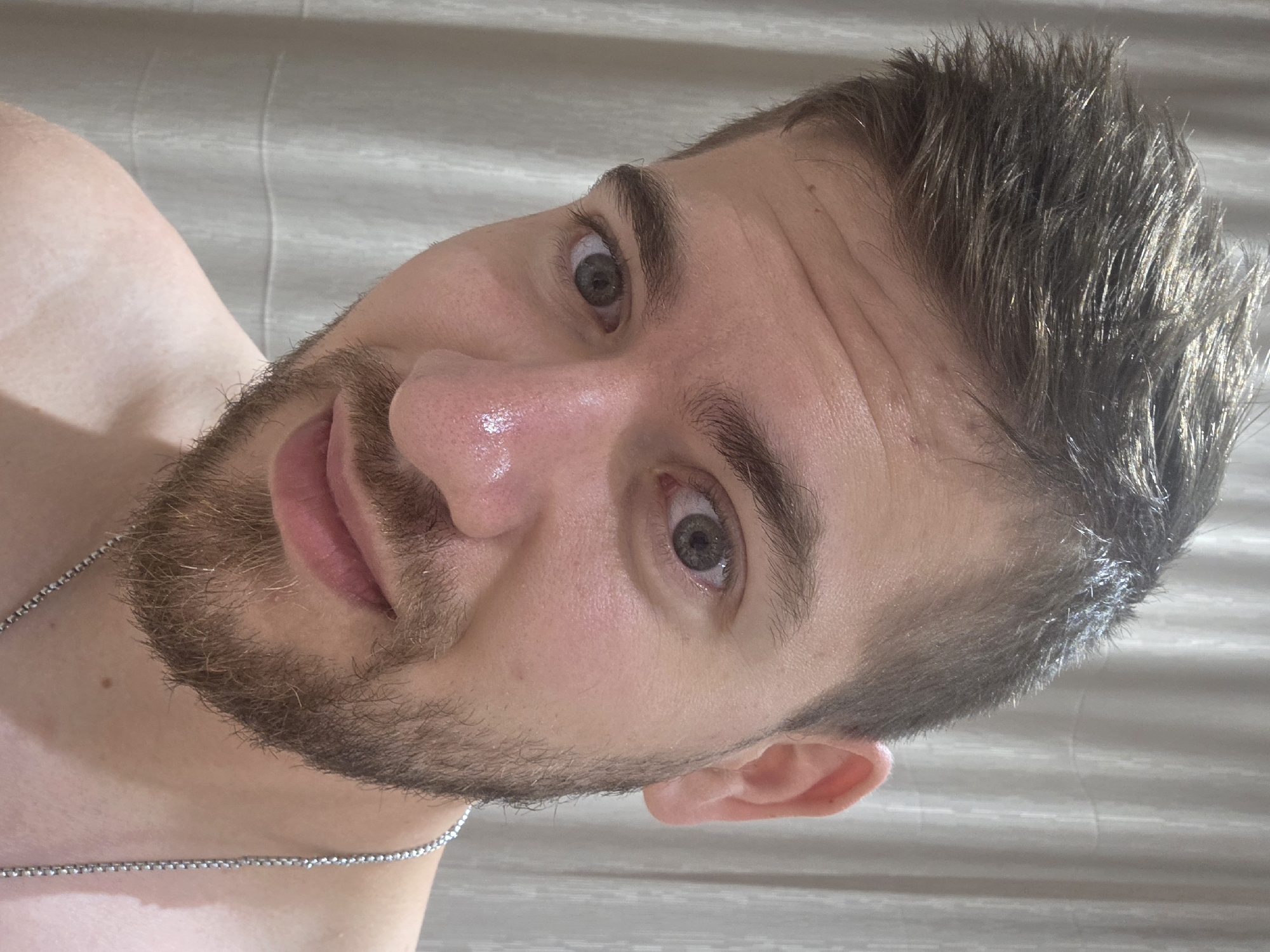 A young, light-skinned man with short, spiky dark hair and a medium-length beard is depicted in a close-up, rotated portrait. He is looking directly at the camera with wide, gray-green eyes and slightly parted lips, suggesting a relaxed or engaging expression. His skin appears healthy, with some natural shine visible on his nose and forehead. He is shirtless, revealing his bare shoulder and chest, where a thin silver chain necklace is visible. A distinct tan line is noticeable across his chest and shoulder. The background is a light, textured surface, possibly a bed sheet or wall, suggesting an indoor setting. There is no visible text in the image. The scene takes place in Yagodovo, Bulgaria.