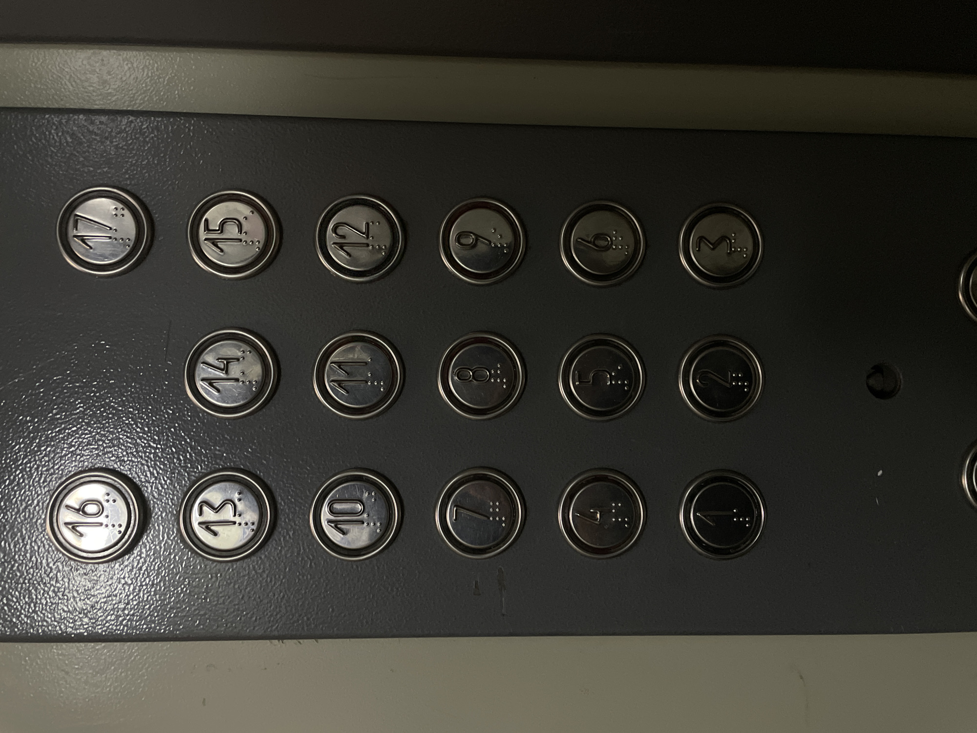 The image shows a close-up of an elevator control panel. The panel is dark gray and has multiple circular buttons arranged in columns. Each button has a number and braille characters embossed on it. The numbers visible are 16, 13, 10, 7, 4, 1, 17, 15, 12, 9, 6, 3, 2, 11, 14, 8, and 5. The panel is mounted on a textured wall, and a portion of a lighter-colored wall or door frame is visible on the left and right edges of the image. There are no people or actions depicted in the image. The scene is an indoor environment, consistent with an elevator lobby or hallway. The location of Rostov-on-Don, Russia, is not evident from the image itself.