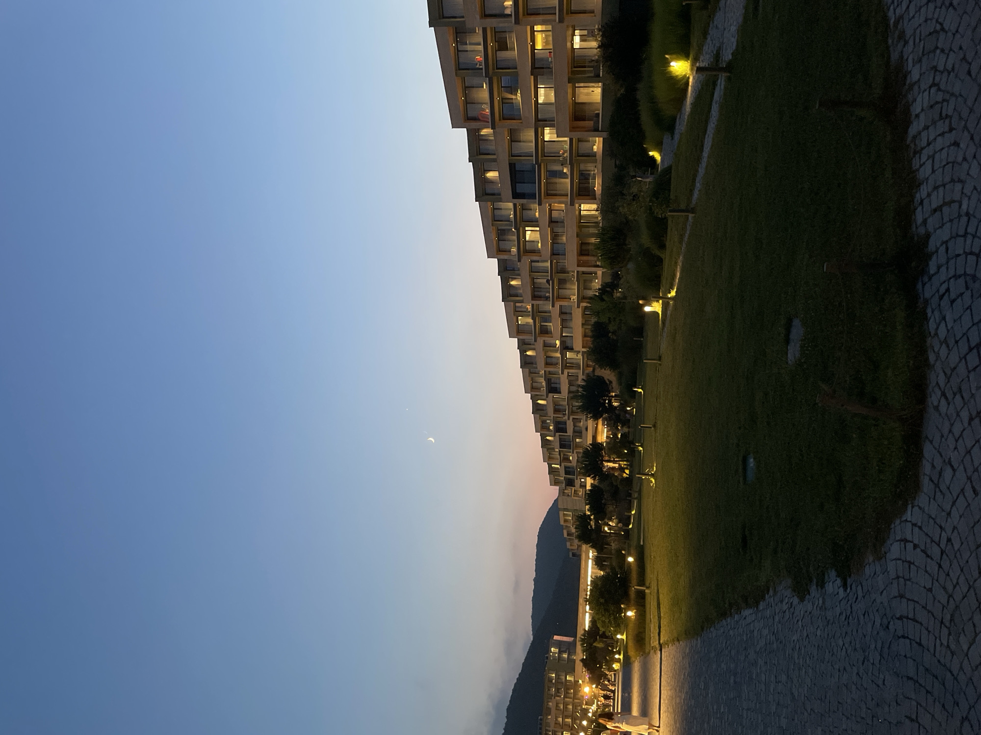 The image depicts a twilight scene in Obzor, Bulgaria, showcasing a modern resort or hotel complex. The architecture features a series of tiered, multi-story buildings with balconies and large windows, illuminated from within, creating a warm glow. In the foreground, a paved walkway, constructed with cobblestones, curves to the left, bordering a well-maintained grassy area dotted with small, yellow pathway lights. Several individuals are visible walking along the pathway, and a lone person stands further down the path, their figure illuminated by the lights. The sky transitions from a pale blue near the horizon, where soft hues of pink and orange are visible, to a deep blue overhead. A crescent moon and a small star are faintly visible in the upper portion of the sky. In the background, dark, silhouetted mountains rise against the twilight sky. The overall atmosphere is serene and suggests an evening ambiance at a resort location.