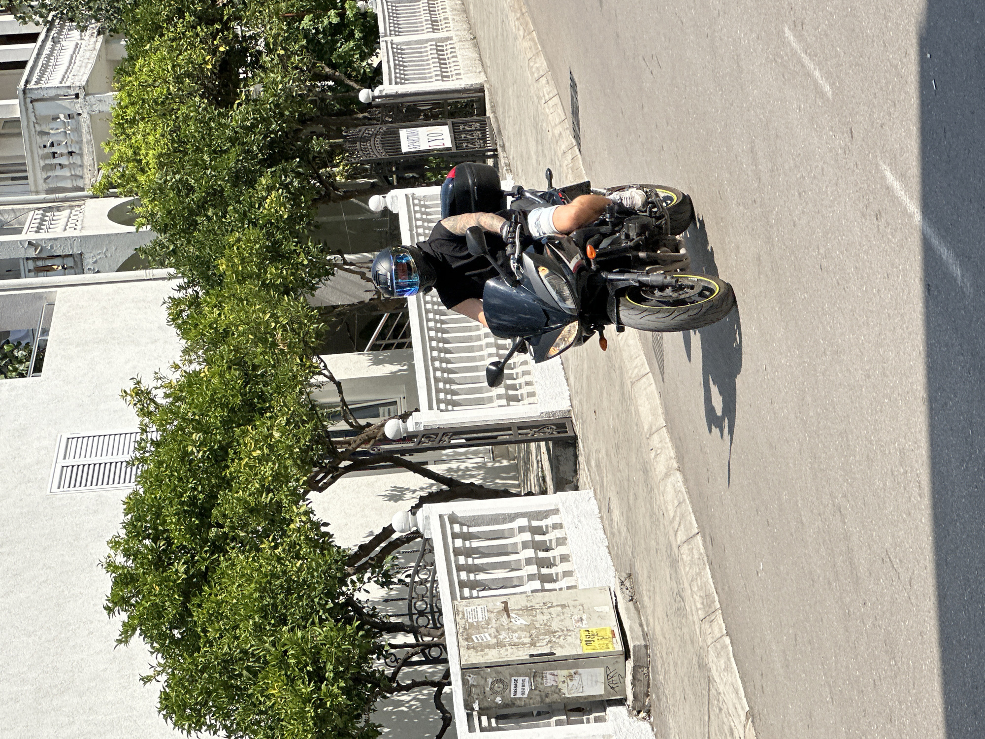 A man wearing a black sleeveless shirt, white shorts, white sneakers, and a black motorcycle helmet with a blue visor rides a black motorcycle with a black top case on a paved road in Budva, Montenegro. He has tattoos on his right arm. The motorcycle has yellow trim on its wheels. The man is moving from the left side of the frame to the right. To the left of the road is a sidewalk with a utility box covered in stickers and graffiti. Behind the utility box and sidewalk is a white building with green trees and a white railing. To the right of the road is another white building with white railings and green trees. A black metal gate with a sign that says "APARTMANI LYO" is visible. The lighting suggests it is a sunny day.