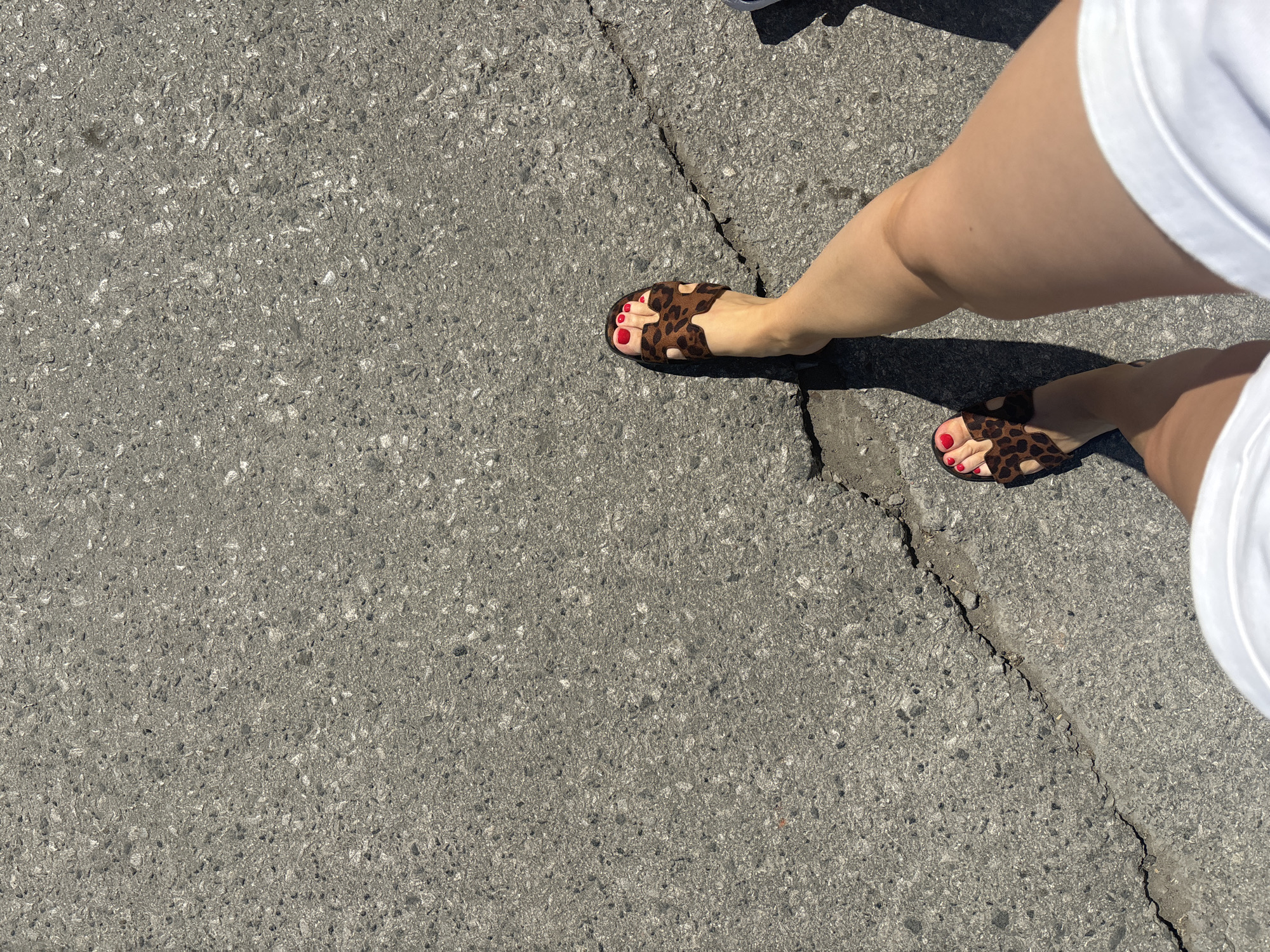 A person stands on a cracked asphalt surface. The person is wearing a pair of brown and black leopard-print sandals with red nail polish on their toes, and white shorts or a skirt. The asphalt has a crack running diagonally across the frame. The photo is taken from above, looking down at the person's legs and feet. The location is unknown, as the city of Perm, Russia is not evident in the image.