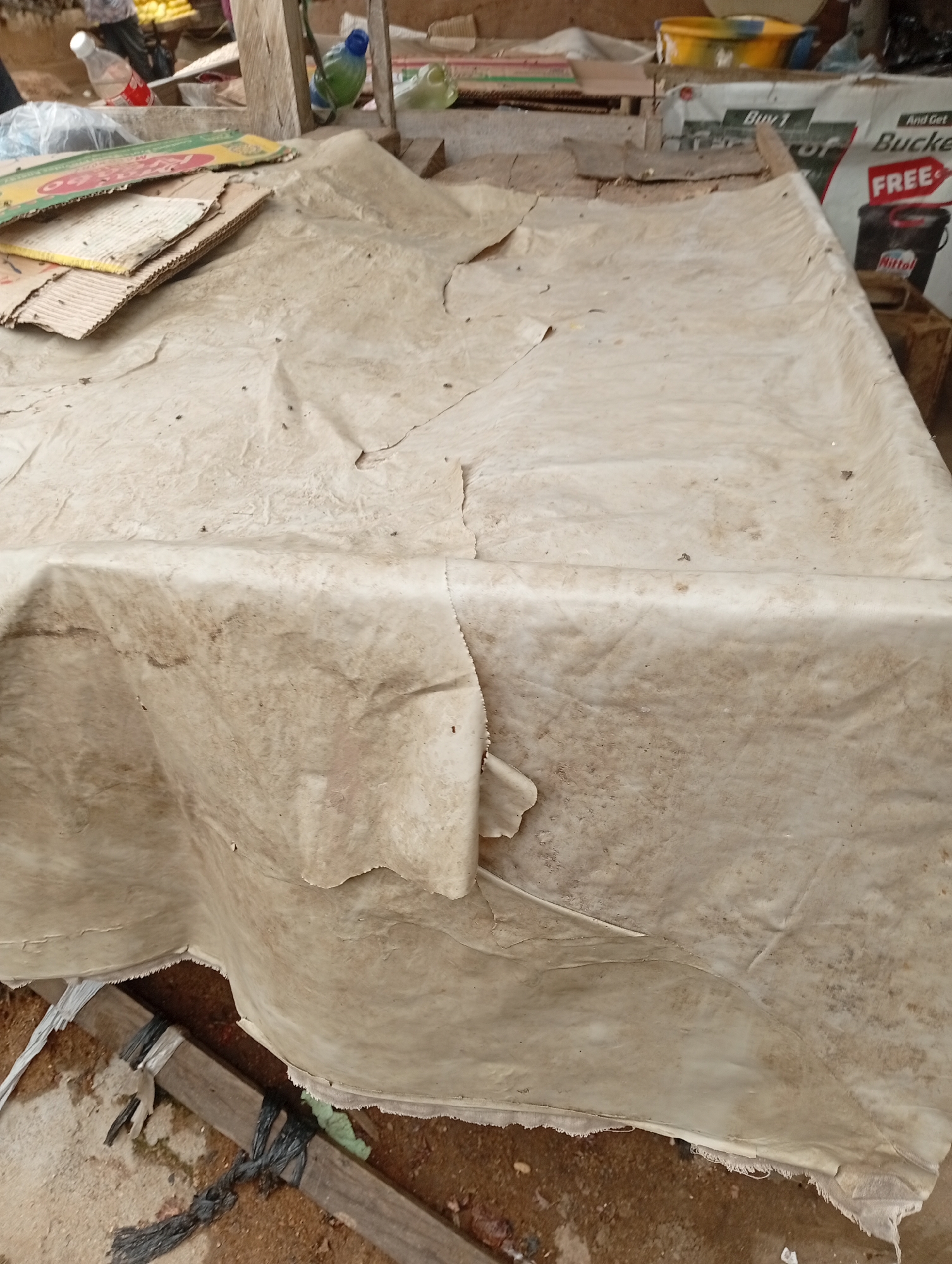 The image captures a close-up view of a covered stall or table, likely in an outdoor market setting in Jalingo, Nigeria. The primary subject is a large, light-colored tarp or sheet that is loosely draped over the structure, creating folds and wrinkles. The material appears worn and stained, with some dirt and debris visible on its surface. In the background, hints of other market elements can be observed, including cardboard boxes stacked to the left and a promotional banner with red lettering visible on the right, suggesting a commercial area. The ground beneath the stall appears to be made of earth and sand. The lighting suggests it is daytime.