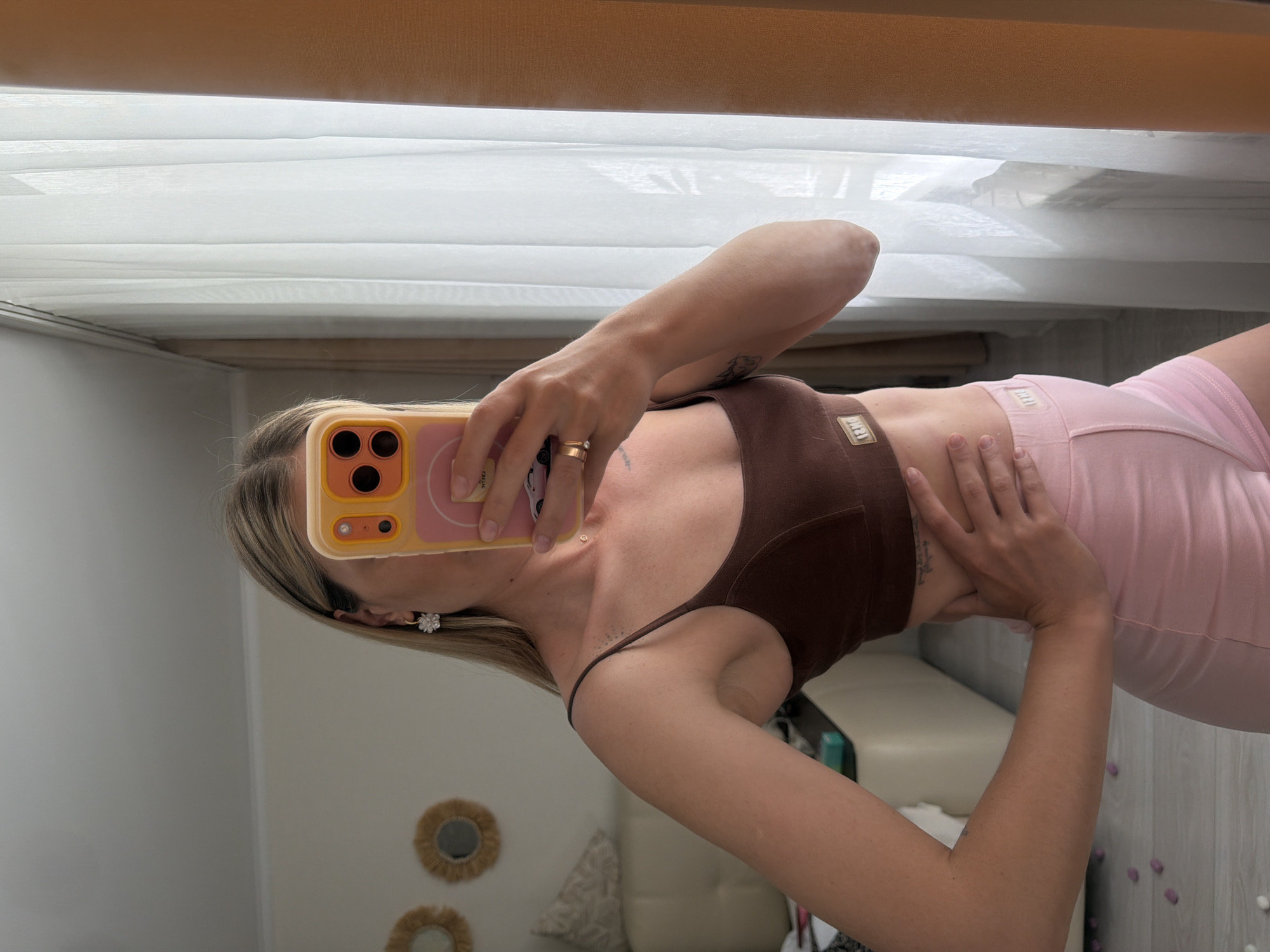 An outdoor, medium shot captures a fair-skinned woman standing in front of a white sheer curtain and a window in what appears to be a residential setting. She is wearing a brown sports bra and pink shorts. The woman is holding up a smartphone with a yellow case to take a mirror selfie. She has blonde hair, an earring in her left ear, and two rings on her right hand. Behind her, to the left, a beige couch and two circular decorative mirrors with rattan frames are visible, along with a reflection of a table and objects on it. The floor is light gray with scattered pink and white decorative items. The scene suggests a personal space, possibly a bedroom or living area, within a home. No elements in the image indicate the city of Semiluki, Russia.