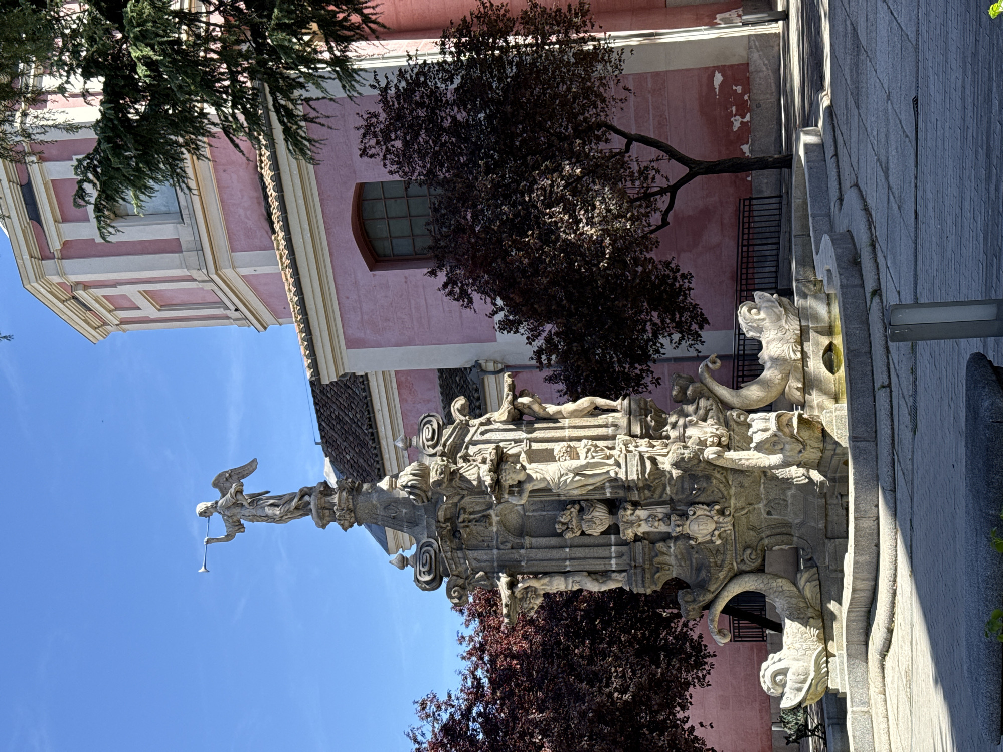 The image shows the Baroque-style Fuente de la Fama fountain in Madrid, Spain, with an angel statue on top playing a trumpet. The fountain is made of stone, adorned with sculptures of mythical creatures, figures, and decorative elements. It is set against a backdrop of a pink building, possibly a church or historical structure, with a tree in the foreground. The sky is bright blue.