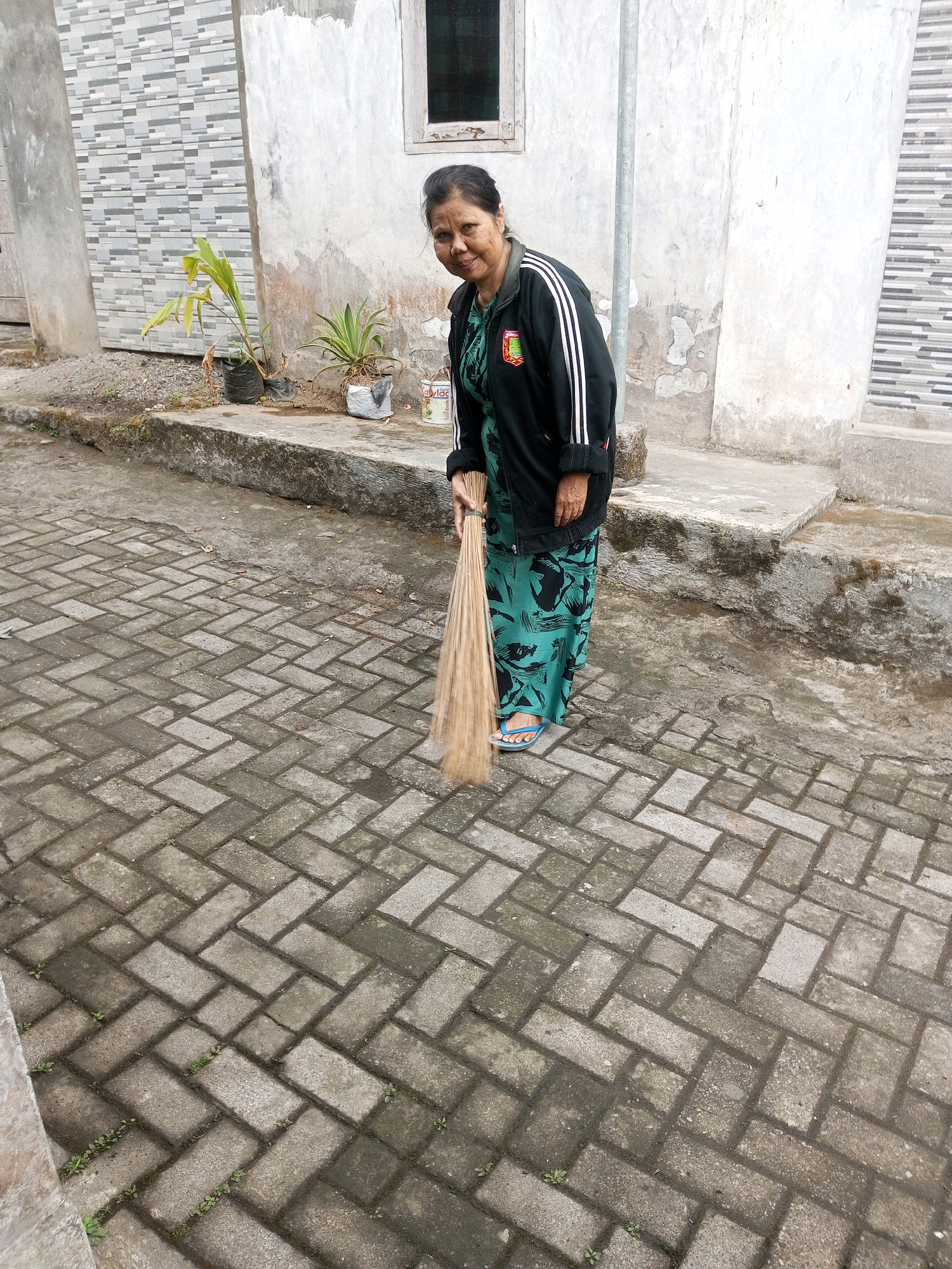 An Indonesian woman stands in what appears to be a residential area in Andongsili, Indonesia, engaged in sweeping the ground with a broom. The woman, wearing a black jacket over a patterned green dress and blue flip-flops, is captured mid-action, with the broom angled towards the ground. Behind her, a building with a white, textured wall and a small window is visible, along with some potted plants. The ground is paved with rectangular interlocking bricks arranged in a herringbone pattern. The overall scene suggests an everyday domestic activity in a local Indonesian setting.