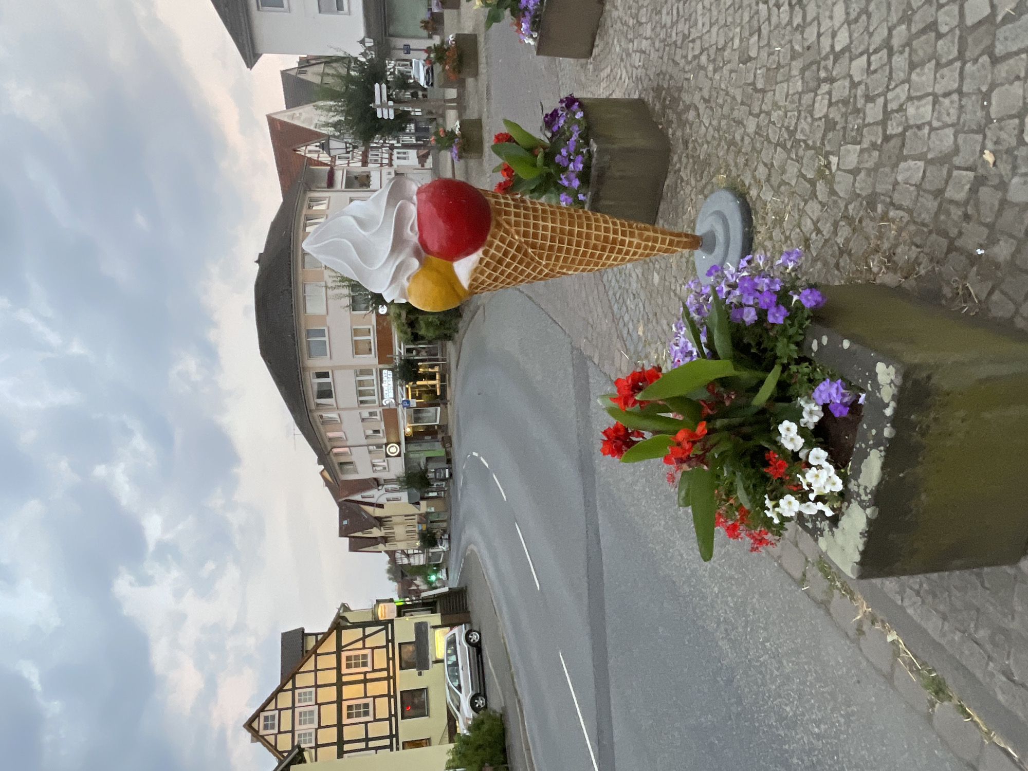 In Neukirchen, Germany, an oversized ice cream cone sculpture stands on a cobblestone sidewalk next to a street. The sculpture features a waffle cone base topped with swirls of white ice cream, two scoops of fruit-flavored ice cream (one yellow, one red), and a red sphere resembling a cherry on top. Two concrete planters filled with red, white, and purple flowers are positioned in front of the ice cream sculpture. Across the street, several buildings with traditional German architecture, including a half-timbered house, line the street. A silver car is parked on the side of the road near the half-timbered building. The sky is overcast with clouds.