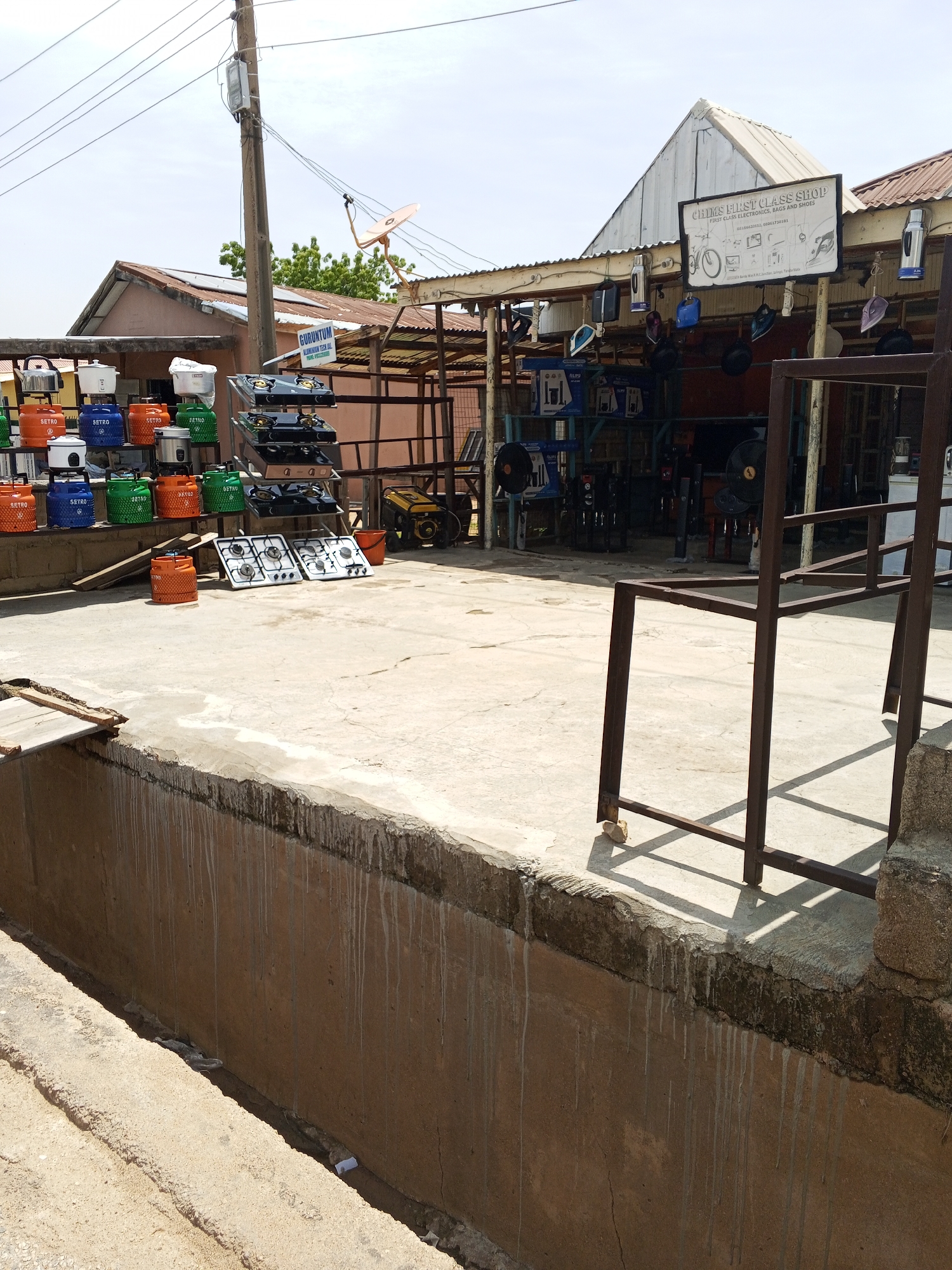 The image depicts an outdoor market scene in Jalingo, Nigeria, showcasing various household appliances and gas cylinders. In the foreground, a concrete platform extends towards the viewer, with a brown, roughly textured wall on the left. To the right, a metal rack structure casts a shadow on the concrete. Behind this, a row of colorful gas cylinders, predominantly orange, blue, and green, are neatly arranged on a raised brick platform. Some cylinders are branded with "SETRO". Above and behind the cylinders, several gas stoves are displayed on a metal rack, with two on a lower shelf and two more stacked above. To the right of the stoves, a yellow generator and a red bucket are visible. The background reveals the facade of a shop with a corrugated metal roof. A prominent sign reading "CHIMS FIRST CLASS SHOP" is mounted on the front of the shop, accompanied by smaller signs and various items hanging from the roofline, including what appear to be electric irons. Further back, in the middle ground, more shop structures and merchandise are visible. Overhead, a telephone pole with electrical wires and a satellite dish extends across the frame, under a bright, partly cloudy sky. The scene appears to be a retail area for home appliances and cooking gas.