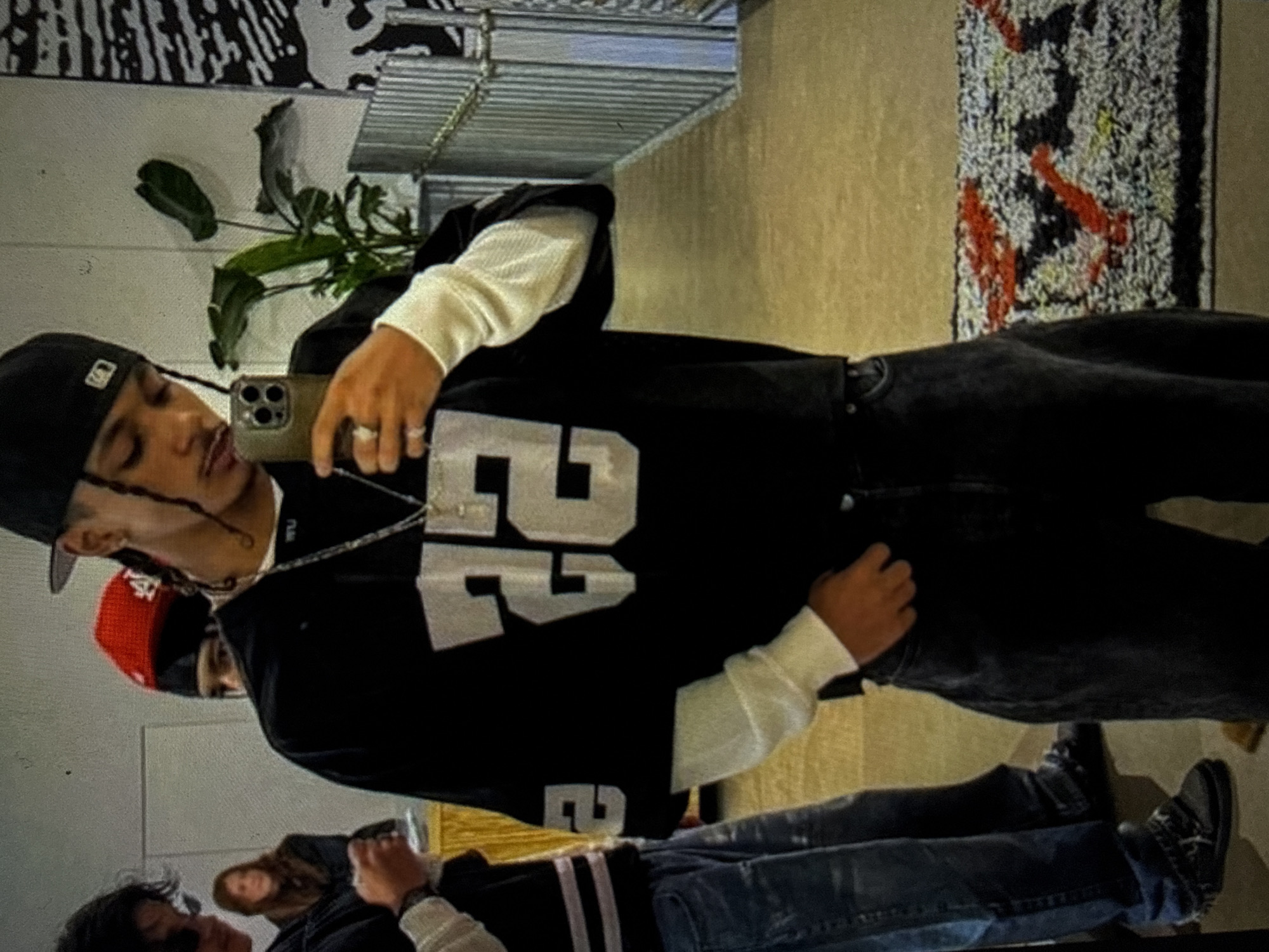 The image captures an indoor scene, identified as being in Kardzhali, Bulgaria. A young male, approximately 20-30 years old, stands centrally, taking a mirror selfie with a dark olive-colored smartphone. He has medium-dark skin, a thin mustache, and dark braided hair or locs, worn with a black backward baseball cap featuring a white square logo. He is dressed in a black short-sleeved jersey-style shirt with large white numbers "22" on the front and a partial "2" on the left sleeve, layered over a white long-sleeved undershirt. A silver chain with a small pendant hangs around his neck, and a ring is visible on his left hand. He wears dark wash jeans.
To the left, a second male individual is partially visible, wearing a dark long-sleeved top with light horizontal stripes on the sleeves and light blue distressed jeans. He has dark shoes with light laces. Further left and out of focus, a female individual with long brown hair is visible, wearing a dark jacket. Behind the second male, the top of a head wearing a bright red knit cap with white lettering is visible, peering from behind.
The environment features white walls. Behind the main subject, a tall green plant in a pot is visible. To the right, a black and white abstract patterned mural or wall art is present, adjacent to a silver metallic slatted or corrugated wall feature that extends into the background. The floor is a light tan or beige color, and a textured rug with abstract patterns in red, black, yellow, and white is visible in the lower right corner. Golden-colored fabric or drapery is faintly visible in the far left background.