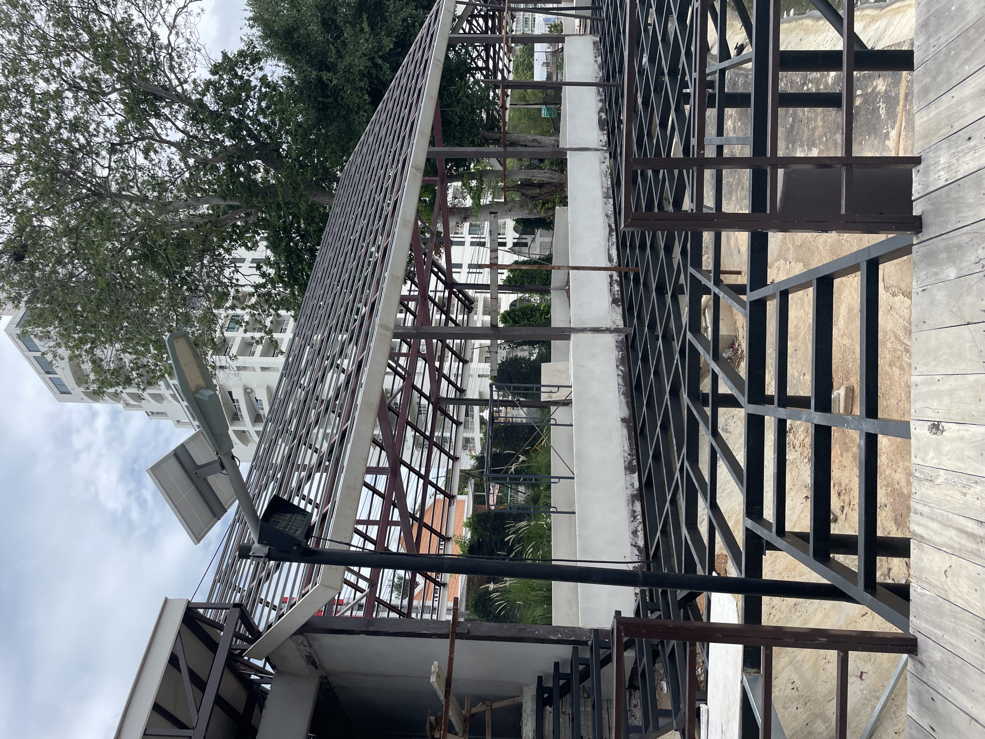 The image depicts a construction site, likely in Pattaya City, Thailand, given the architectural style of the buildings in the background and the tropical vegetation. In the foreground, a weathered wooden walkway leads to a partially constructed building. The building itself is composed of exposed steel framing, with a partially completed roof structure. A solar-powered street lamp is mounted on a pole near the roof's edge. Beyond the construction site, a lush green tree stands prominently, and in the distance, a multi-story white building with numerous balconies suggests an urban or resort setting. The sky is overcast, indicating a cloudy day. There are no people visible in the image. The scene is one of ongoing development, with the raw structural elements of the building contrasting with the natural elements and established architecture in the background.