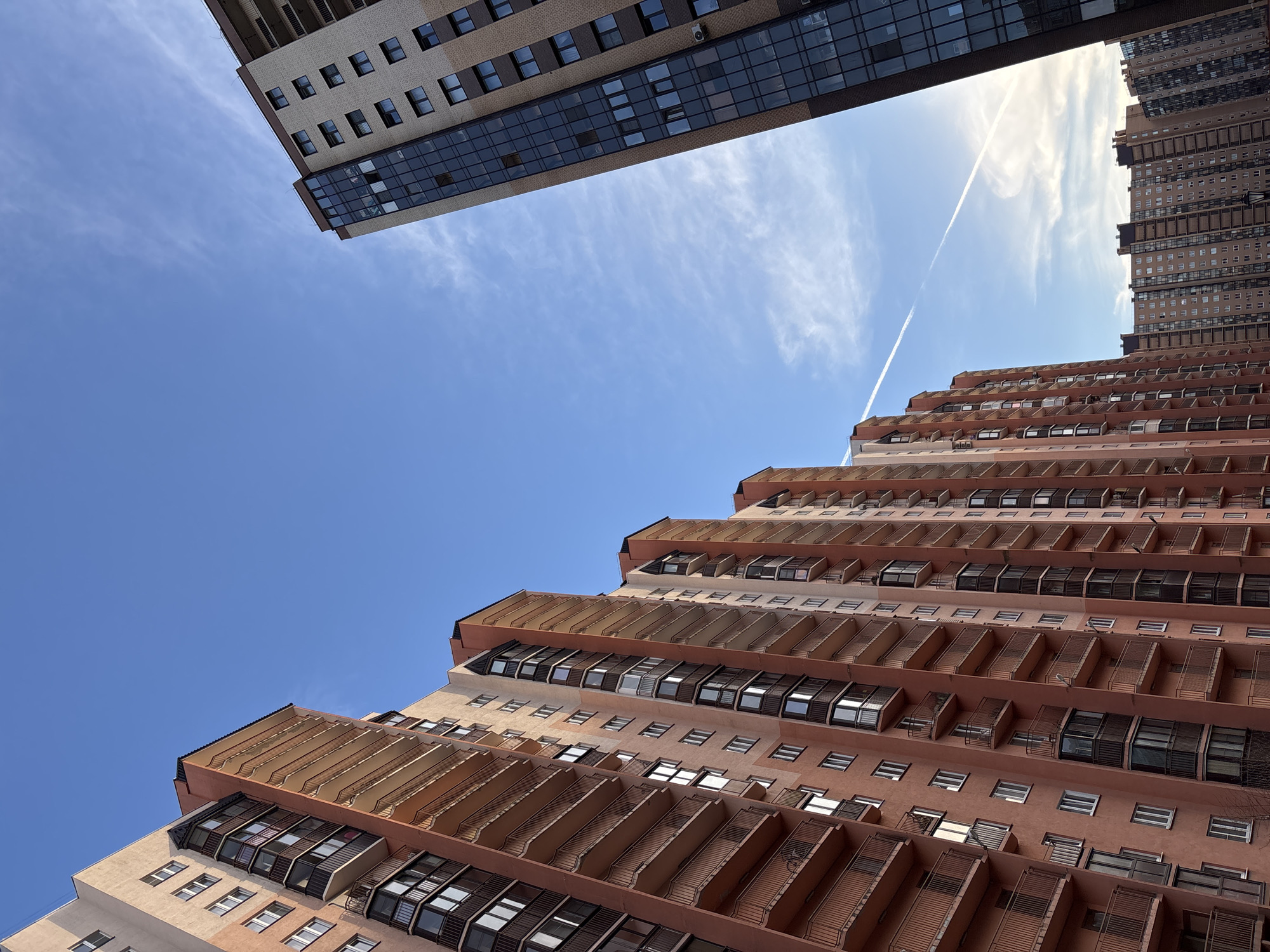 The photograph shows a view looking up at two high-rise buildings in Kudrovo, Russia, against a clear blue sky with a contrail. The building on the right is a stepped, salmon-colored apartment building with repeating patterns of windows and balconies. The building on the left is a taller, rectangular, modern structure with a glass facade.