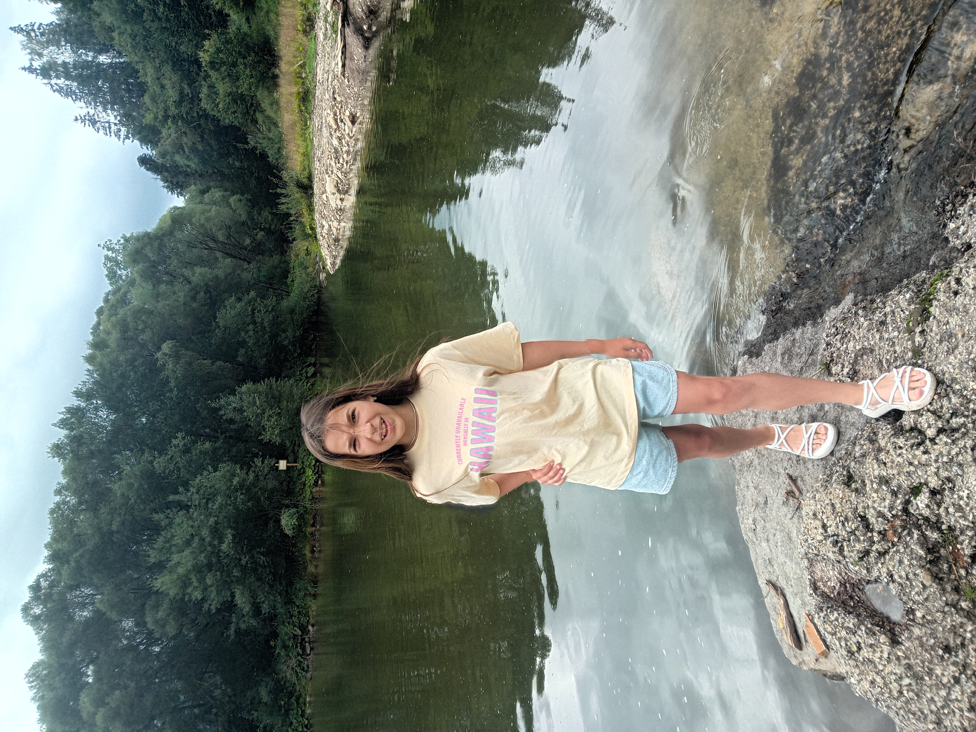 A young girl with long brown hair is standing on the rocky bank of a river, with a forest visible in the background. She is wearing a light yellow t-shirt with the text "CURRENTLY UNAVAILABLE MENTALLY IN HAWAII" and light blue shorts. She is also wearing white sandals. The river has calm water with reflections of the trees and sky. The overall scene appears to be a natural outdoor setting, possibly in the outskirts of Habovka, Slovakia, given the lush greenery and river.