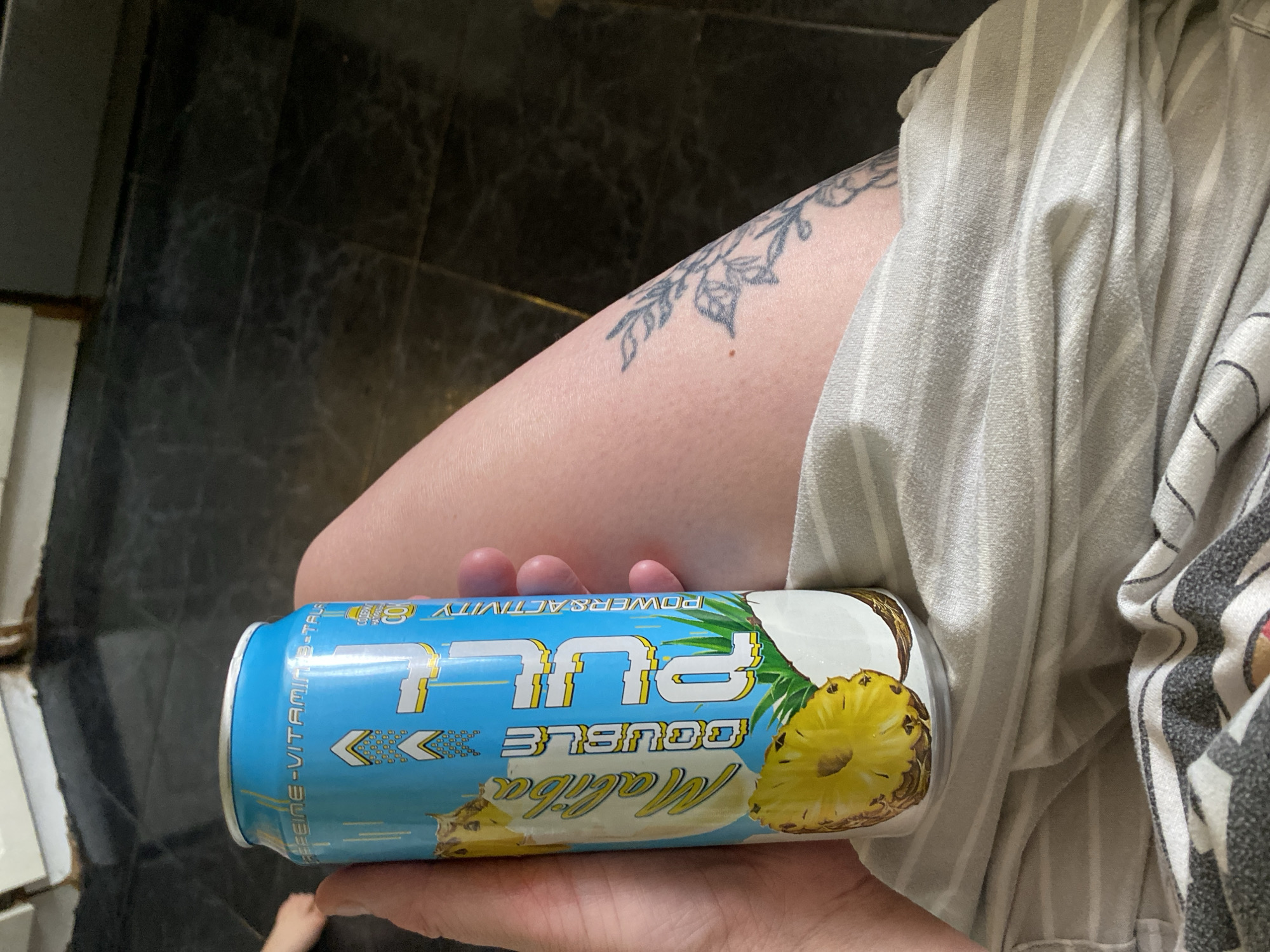 A hand holds a can of "Maliba Double Pull" energy drink, pineapple and coconut flavor. The person is wearing a striped garment and has a floral tattoo on their leg. The background shows dark tiled flooring and a glimpse of white cabinetry. The location is unknown and cannot be identified as Chaikovsky, Russia from the provided image.