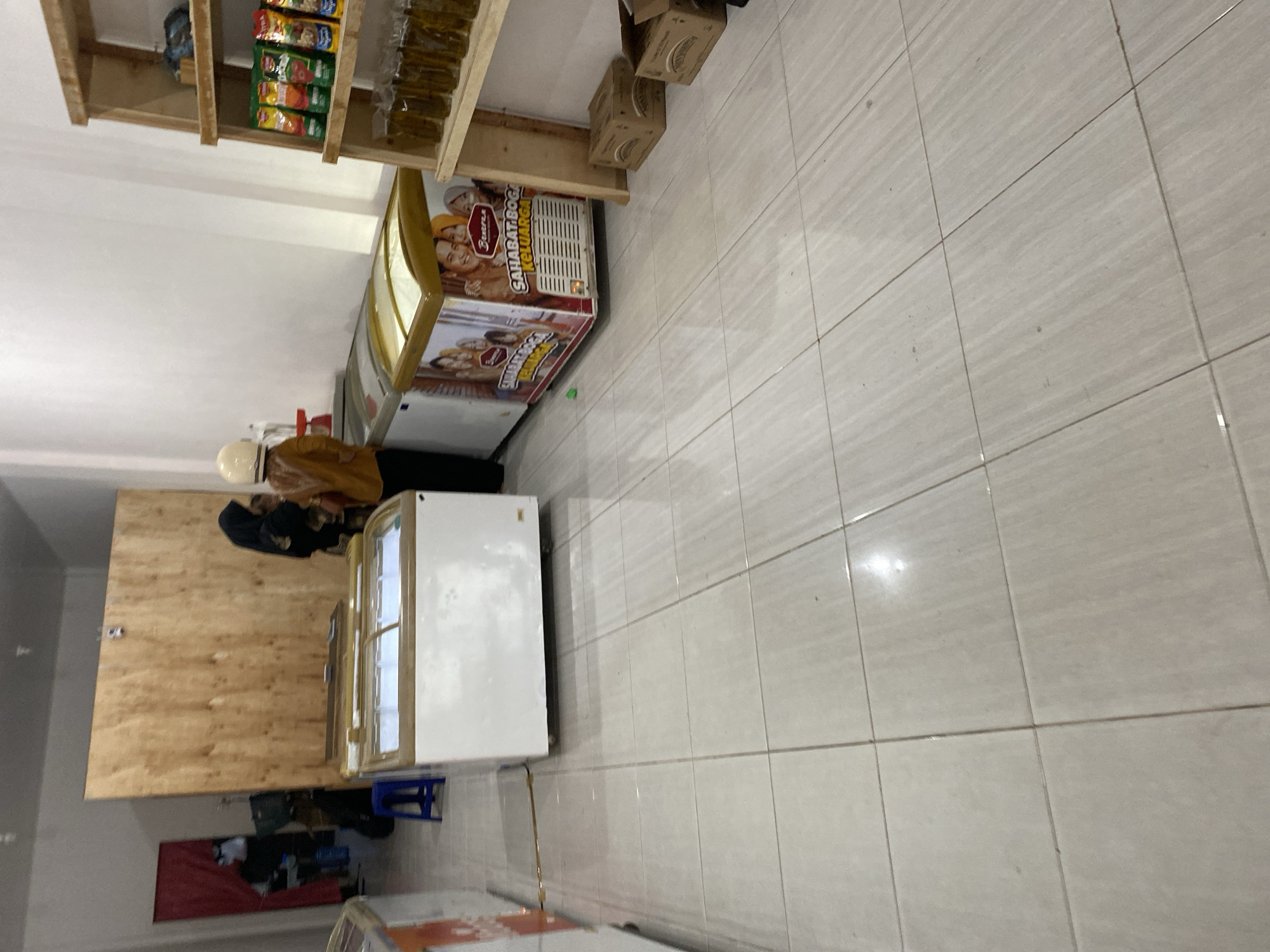 In the city of Payatieng, Indonesia, an indoor scene captures a retail environment. The focal point is a row of chest freezers, with two prominent white models on the left and a decorated one featuring an image of a family and "SAHABAT BOGA KELUARGA" text on the right. Behind the freezers, a large wooden panel forms a backdrop.
Two individuals are present. On the right, a person wearing a yellow top and a white helmet stands facing away from the camera. To their left, another person is partially visible, wearing a black hijab and dark clothing. A small blue stool is situated near the left freezer.
To the right of the freezers, a wooden shelf unit displays various products. The top shelf holds several small packets of food items, appearing to be snacks or sauces, in colorful packaging. Below this, a row of glass bottles filled with a yellowish liquid, likely oil, are neatly arranged. At the base of the shelf unit, two cardboard boxes are placed on the floor.
The floor is tiled with light-colored square tiles, showing some subtle variations in pattern and a few scattered dark specks. The overall lighting suggests an indoor commercial space.