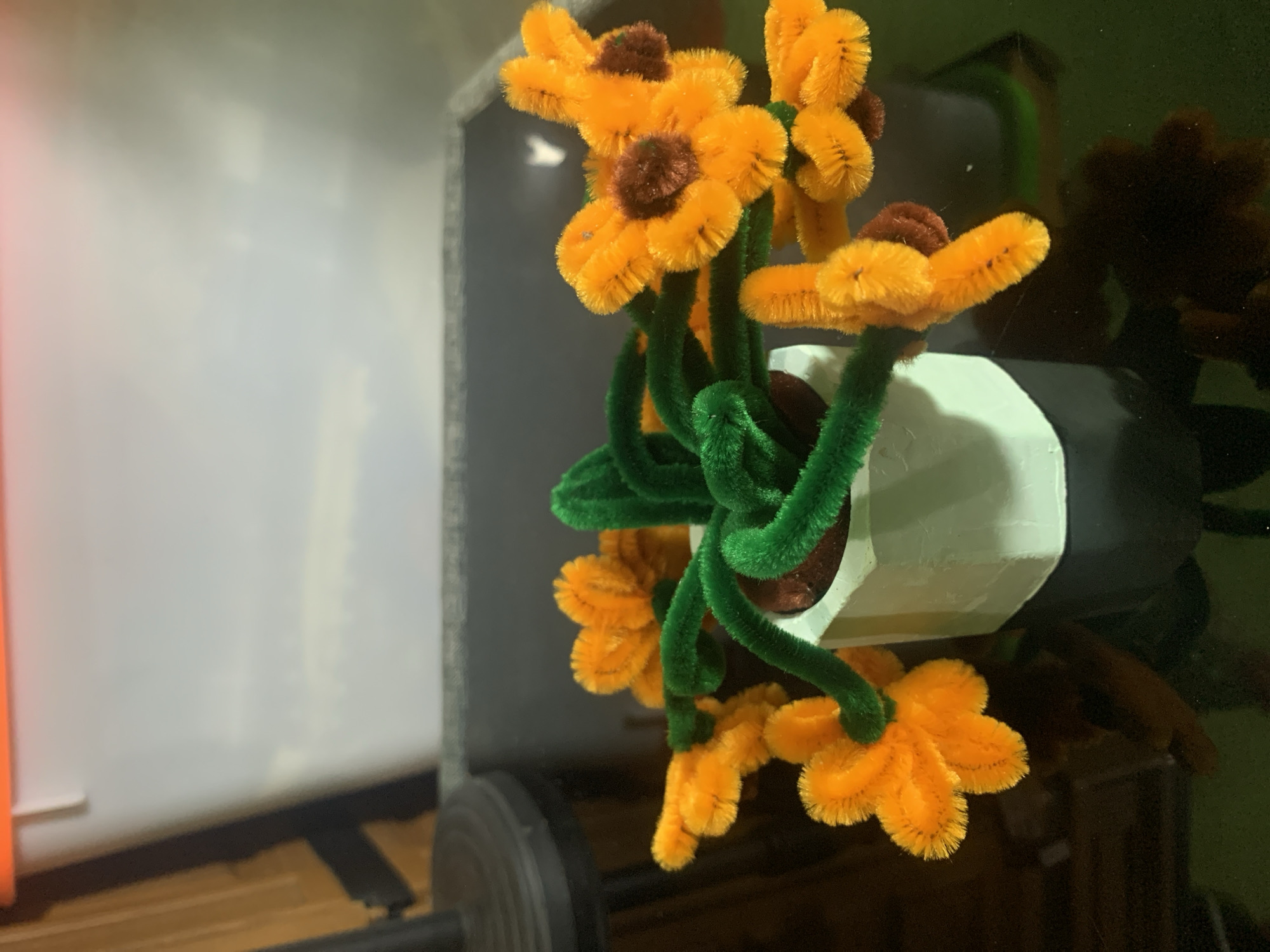 This is a close-up, eye-level shot of a craft project. In the foreground, a small, light green, hexagonal pot holds a bundle of fluffy green pipe cleaners, twisted to resemble stems. Attached to these stems are several bright orange, fluffy pipe cleaner flowers, some with dark brown centers. The pot and flowers are resting on a dark, reflective surface, possibly a table. In the blurred background, there appears to be a white and orange rectangular object. The image does not contain any elements that would place it in Kuala Pembuang, Indonesia or any specific city.