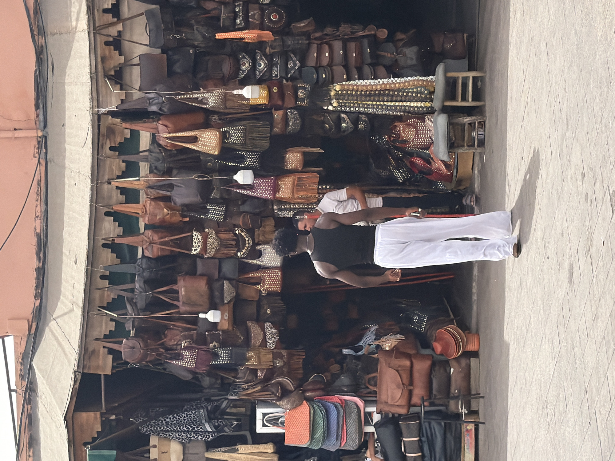 In Marrakesh, Morocco, a young Black man walks away from the camera towards a market stall brimming with an array of leather bags and accessories. He is wearing a black tank top and flowing white pants. Behind him, a second man, wearing a white short-sleeved shirt, is partially visible, engaging with the stall owner or customer. The stall displays a dense collection of handbags of various sizes, colors, and styles, many adorned with intricate beadwork, studs, or fringe. Woven baskets and stacked hats are also visible. The stall is covered by a striped awning, typical of the Djemaa el-Fna square in Marrakech, suggesting this is a scene from one of its bustling souks. The ground is paved with light-colored tiles, and shadows indicate a bright, sunny day.