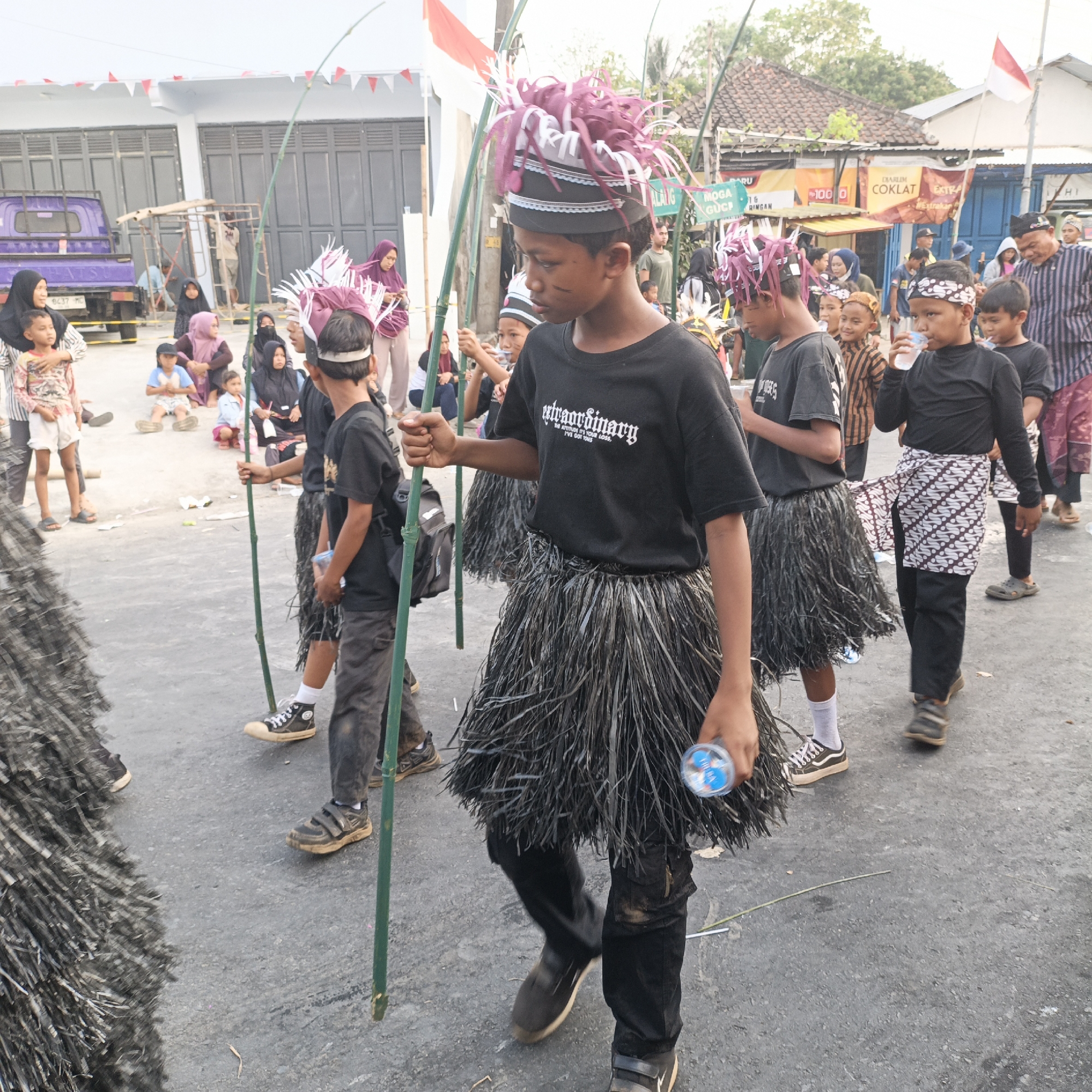 The image captures a street scene in Randudongkal, Indonesia, likely during a cultural event or parade. Several young boys, dressed in traditional attire including feathered headdresses and raffia skirts, are walking in procession, each holding a long bamboo stick. One boy in the foreground wears a black t-shirt with white text that reads "Extraordinary" and a patterned sash over dark trousers. Another boy is seen drinking from a plastic bottle. Adults and children are gathered on the sides, observing the procession. In the background, buildings with visible signs and a purple truck add to the urban setting. Red and white flags are strung along the street, suggesting a patriotic or celebratory occasion. The ground is paved with asphalt, and there is debris scattered across the surface.