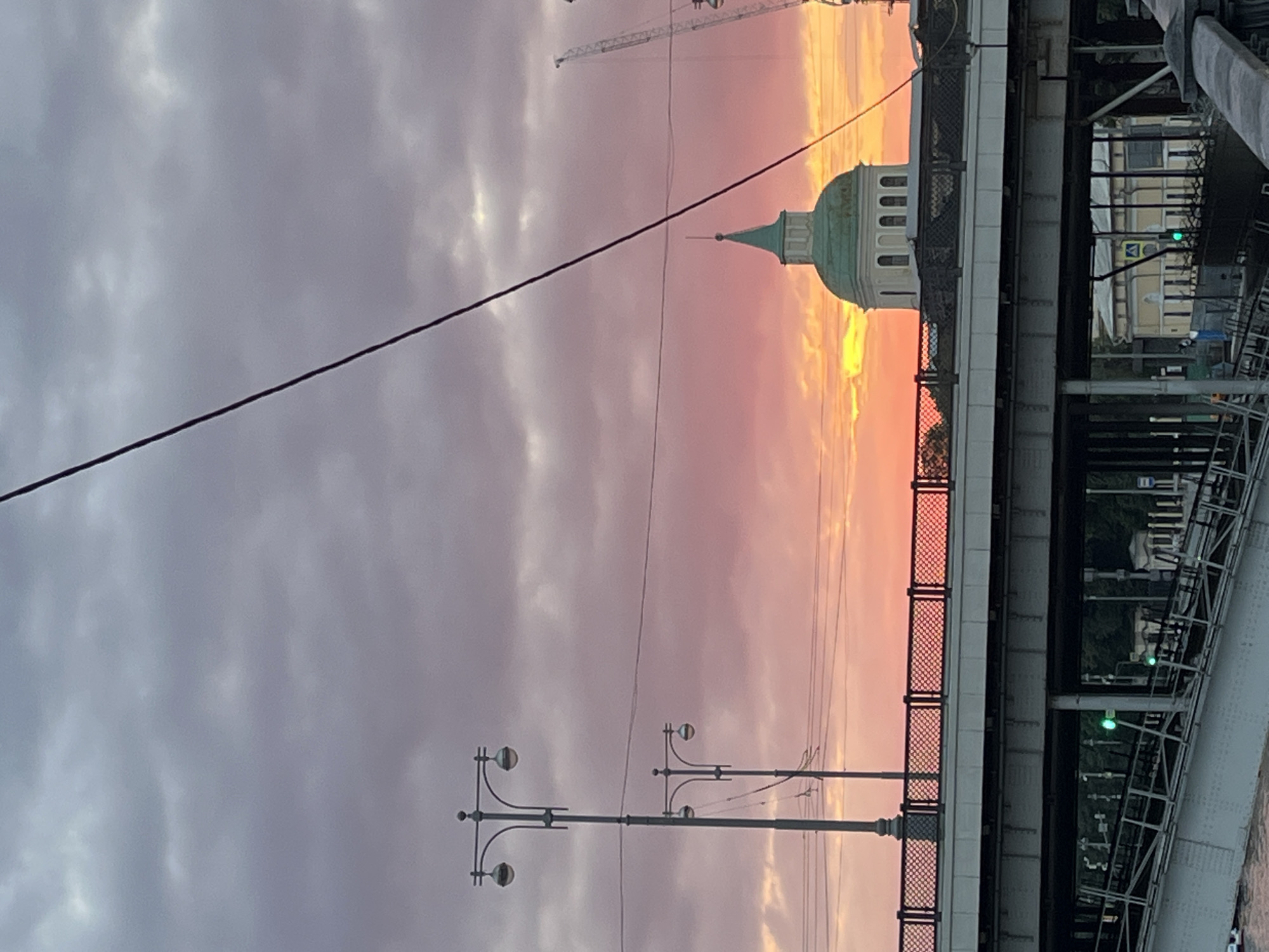 The image shows a sunset over a city, likely Moscow, Russia, as evidenced by the architectural style and the presence of a domed building resembling a church or observatory, possibly part of the Moscow State University complex or another historical building. The sky is filled with dramatic clouds, painted in shades of purple, pink, and orange, with the sun breaking through near the horizon. A bridge spans across the foreground, with its metal structure and railings visible. On the bridge, streetlights with multiple globes are positioned, adding to the urban atmosphere. In the background, several construction cranes are visible, indicating ongoing development in the city. The lower part of the image shows the underside of the bridge, revealing more of its structural components and the water below, suggesting it crosses a river. The scene is devoid of any people.