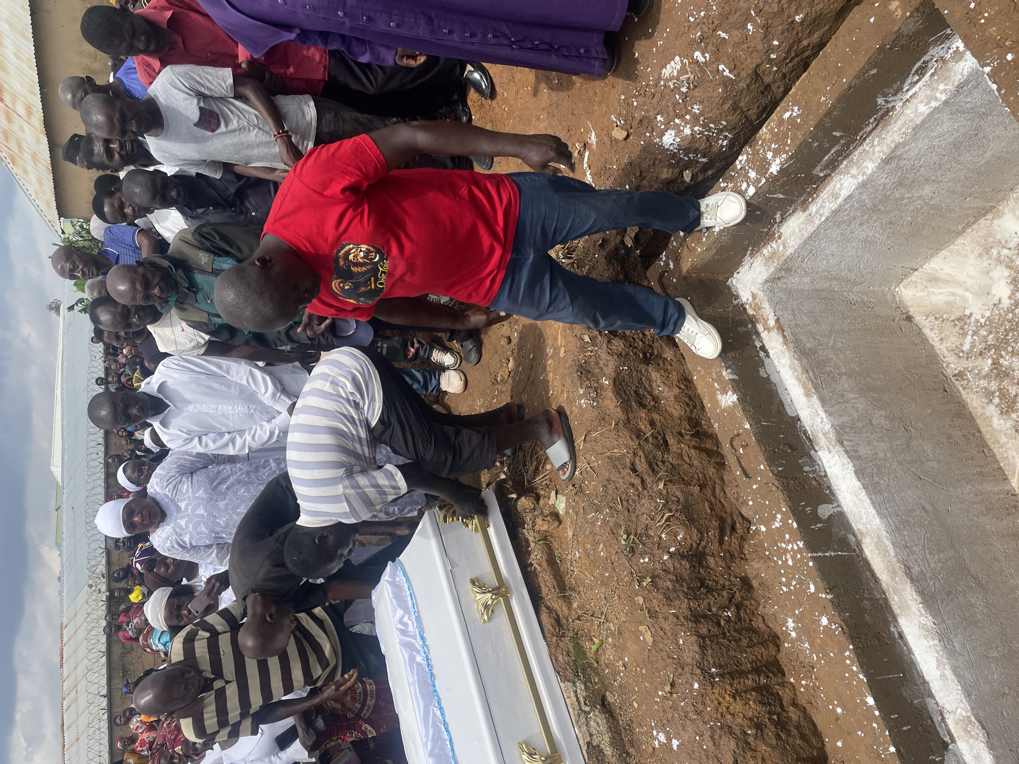 At a burial in Gura Top, Nigeria, a crowd of mourners gathers around an open grave containing a white coffin with gold trim. Men in casual attire and some in religious robes stand on the edge of the grave, while others are seen in the background. The ground is dry earth, with the grave dug into the soil, revealing a concrete lining. Some of the attendees are looking towards the coffin, while others are engaged in conversation or observing the proceedings. The sky above is overcast.
