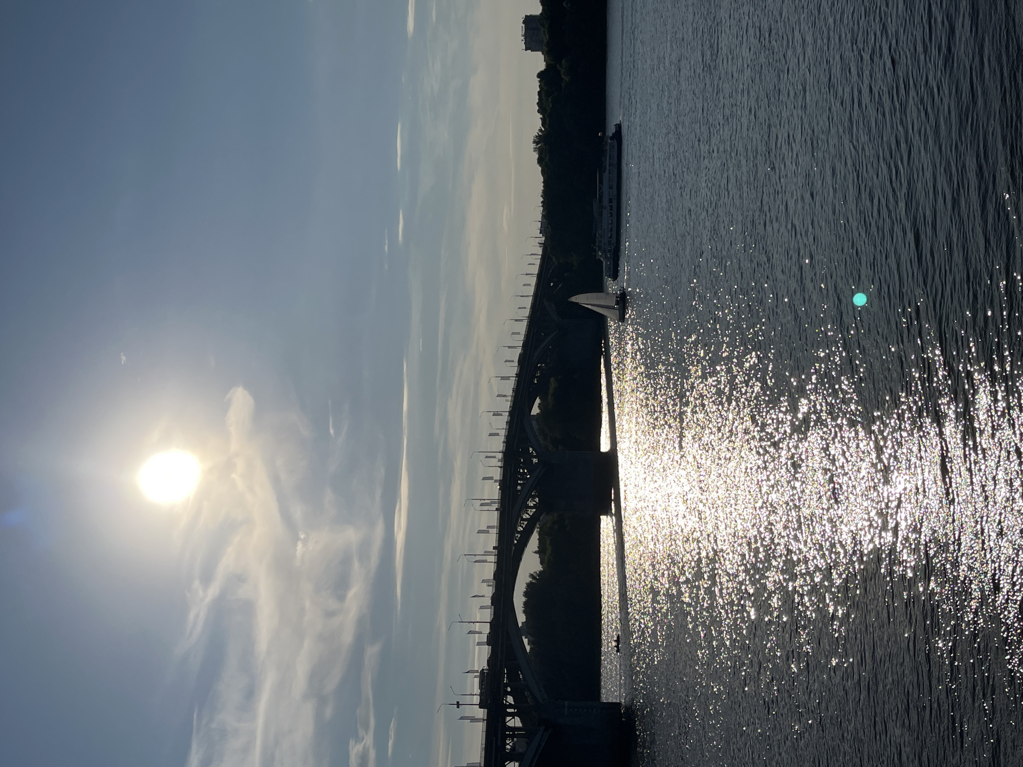 The image depicts a wide river in Nizhny Novgorod, Russia, under a bright, hazy sun in a blue sky with scattered clouds. A large arched bridge spans the river in the mid-ground, with vehicles and streetlights visible on its deck. The water's surface is brilliantly illuminated by the sun, creating a shimmering, sparkling effect. Two boats are present on the water: a small sailboat with a white sail to the left of center, and a larger passenger boat behind it. On the far bank, a dense line of trees is visible, with a few buildings peeking out above them. The overall atmosphere is one of a serene summer day.
