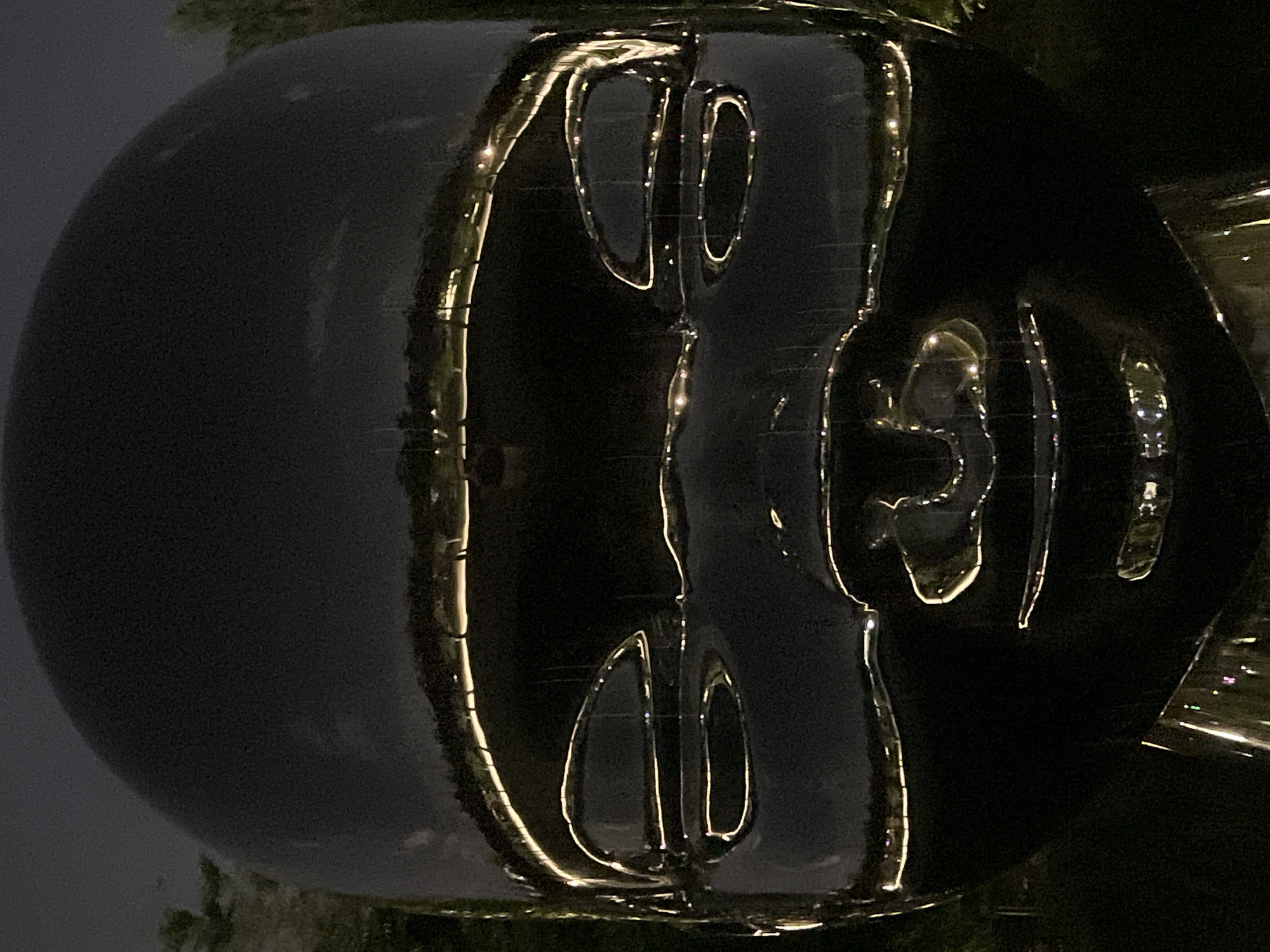 The image is a close-up, front-facing shot of a large, dark, reflective sculpture of a baby's head at night. The sculpture is highly polished and reflects the surrounding environment, including lights and trees. The reflective surface creates distorted images of what appears to be a park or outdoor area with a pathway illuminated by a string of lights. The sculpture's features, such as the eyes and mouth, are exaggerated and stylized, appearing as smooth indentations within the reflective surface. The overall impression is one of a modern, abstract art installation. The location is not specified in the provided information.