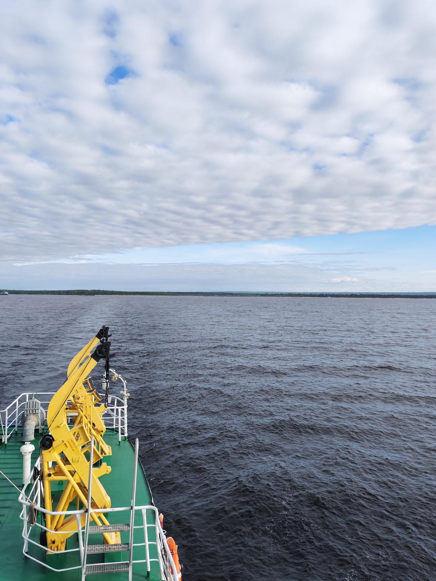 A view from a boat in Ozernoe Ust'e, Russia shows a partially cloudy sky above a large body of water. A green and white boat deck with a yellow crane is visible in the foreground. Across the water, a thin strip of trees lines the horizon.